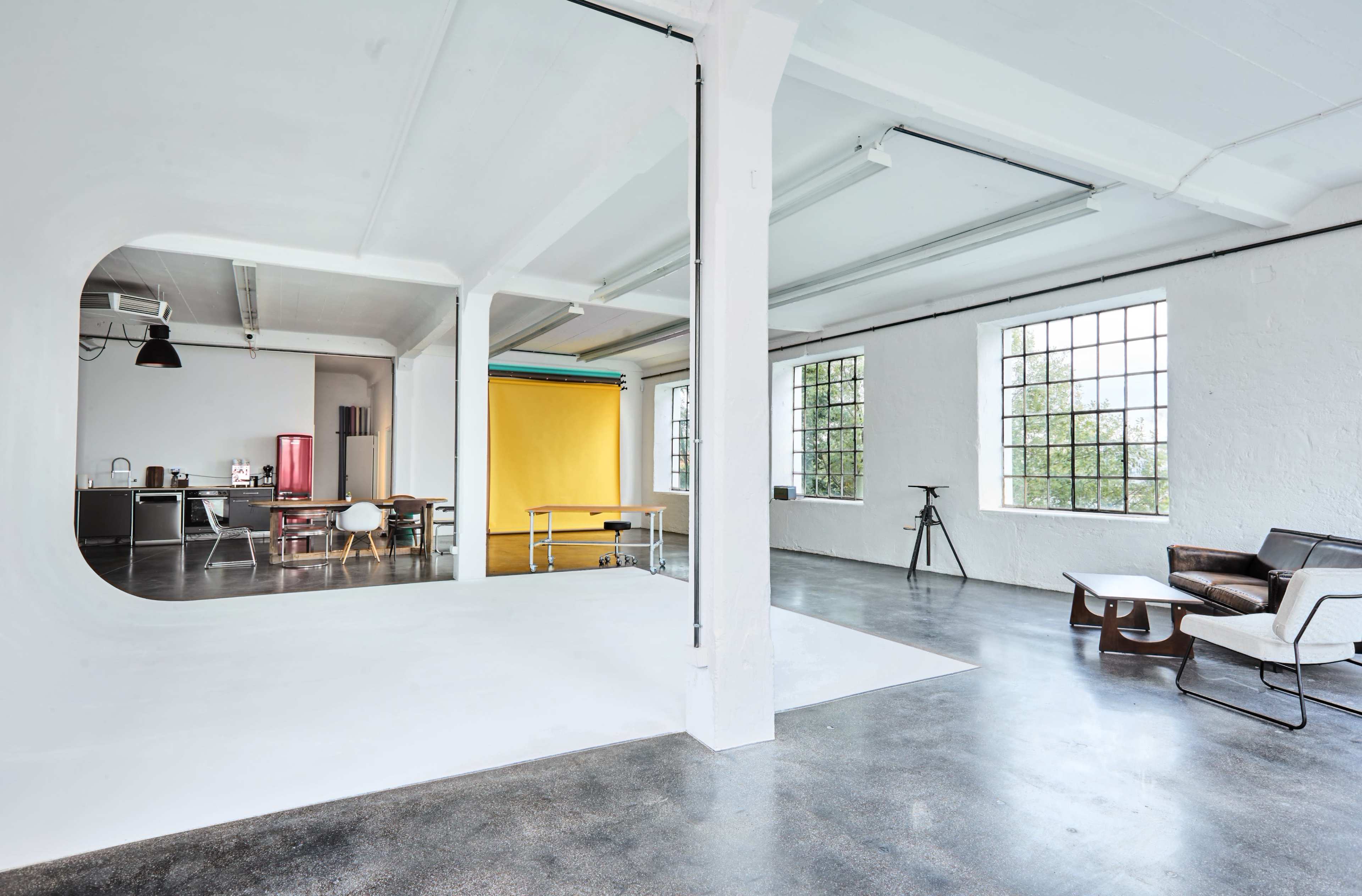 The image shows a spacious, well-lit studio with a white wall, a modern seating area, and a kitchen space visible in the background.