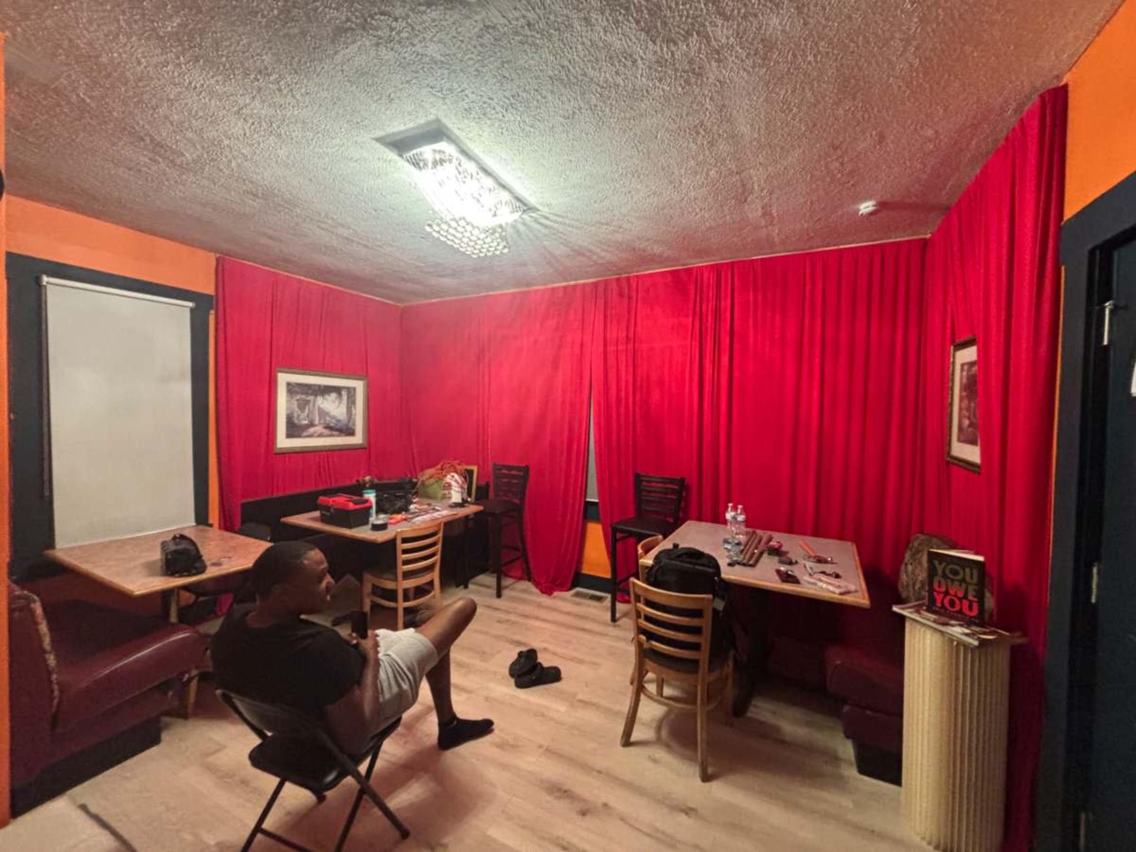 The image shows a small room with red curtains and walls, featuring two tables, chairs, and scattered personal items.