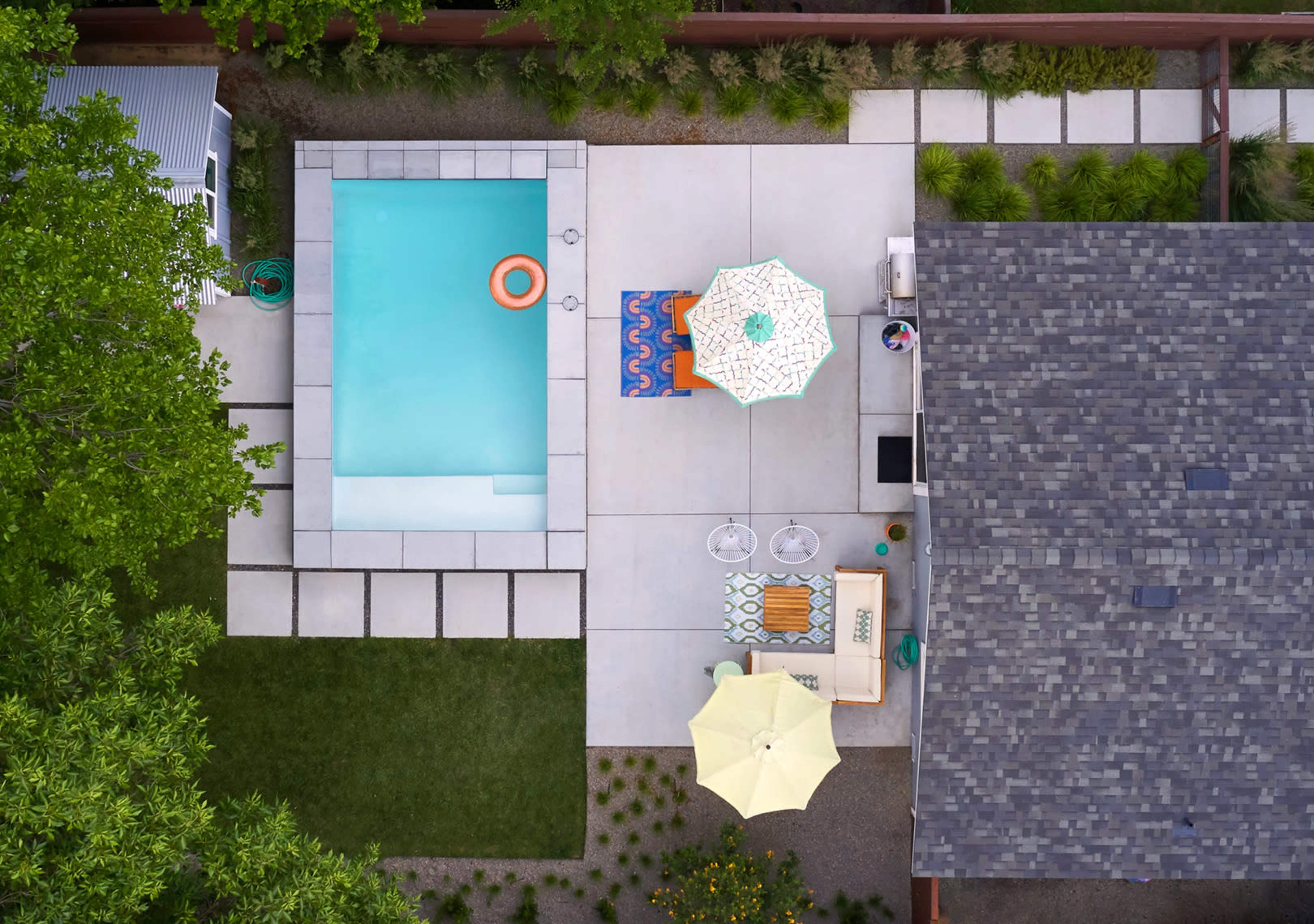 The image shows an aerial view of a backyard featuring a rectangular swimming pool, outdoor furniture, and umbrellas on a concrete patio beside a house.