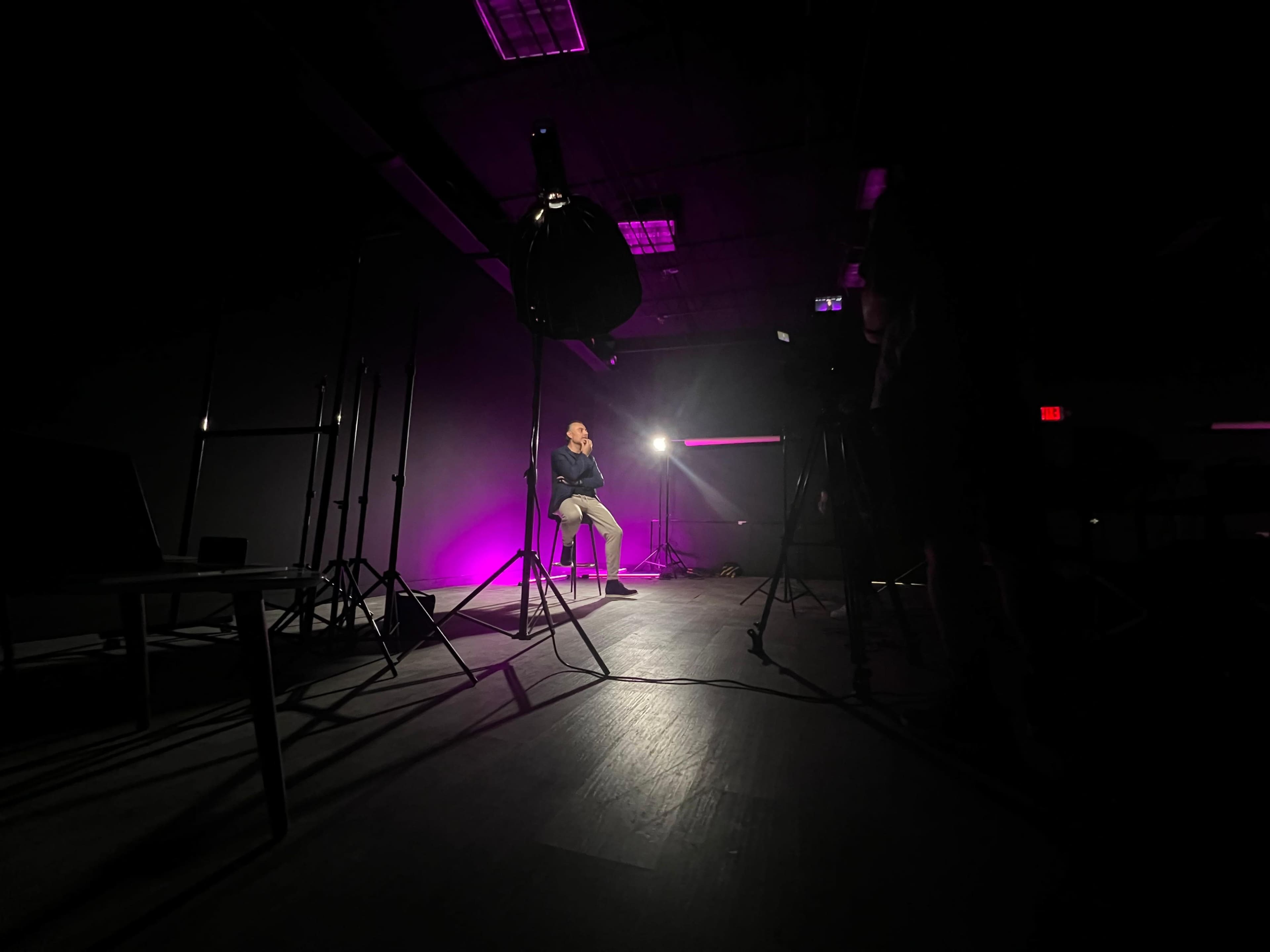 A person sits on a stool in a dimly lit studio, illuminated by a bright light with purple hues and surrounded by camera equipment and stands.