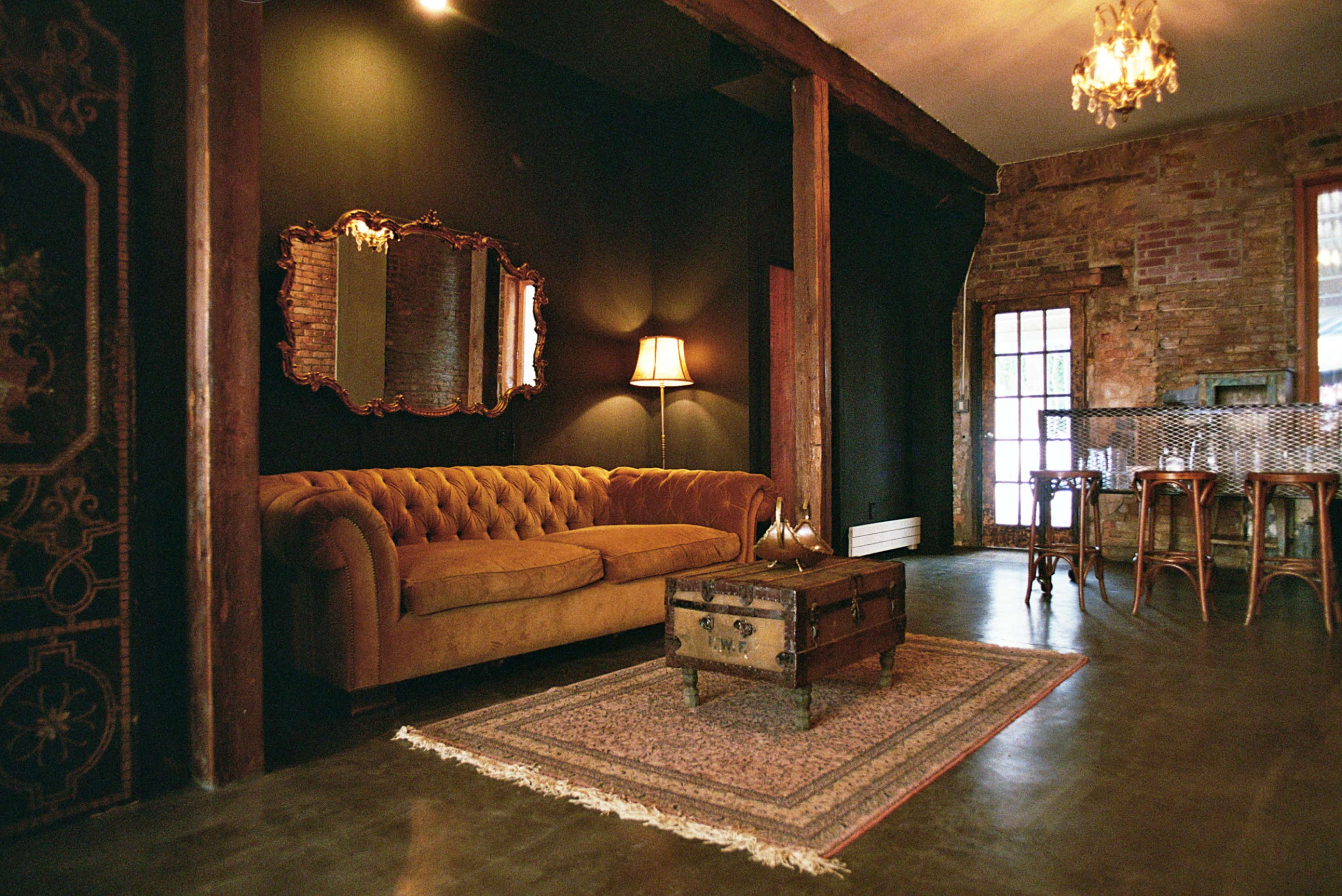 The image shows a cozy living space featuring a tufted brown sofa, a vintage-style mirror, a wooden coffee table, and a bar area with stools against a wall of exposed bricks.