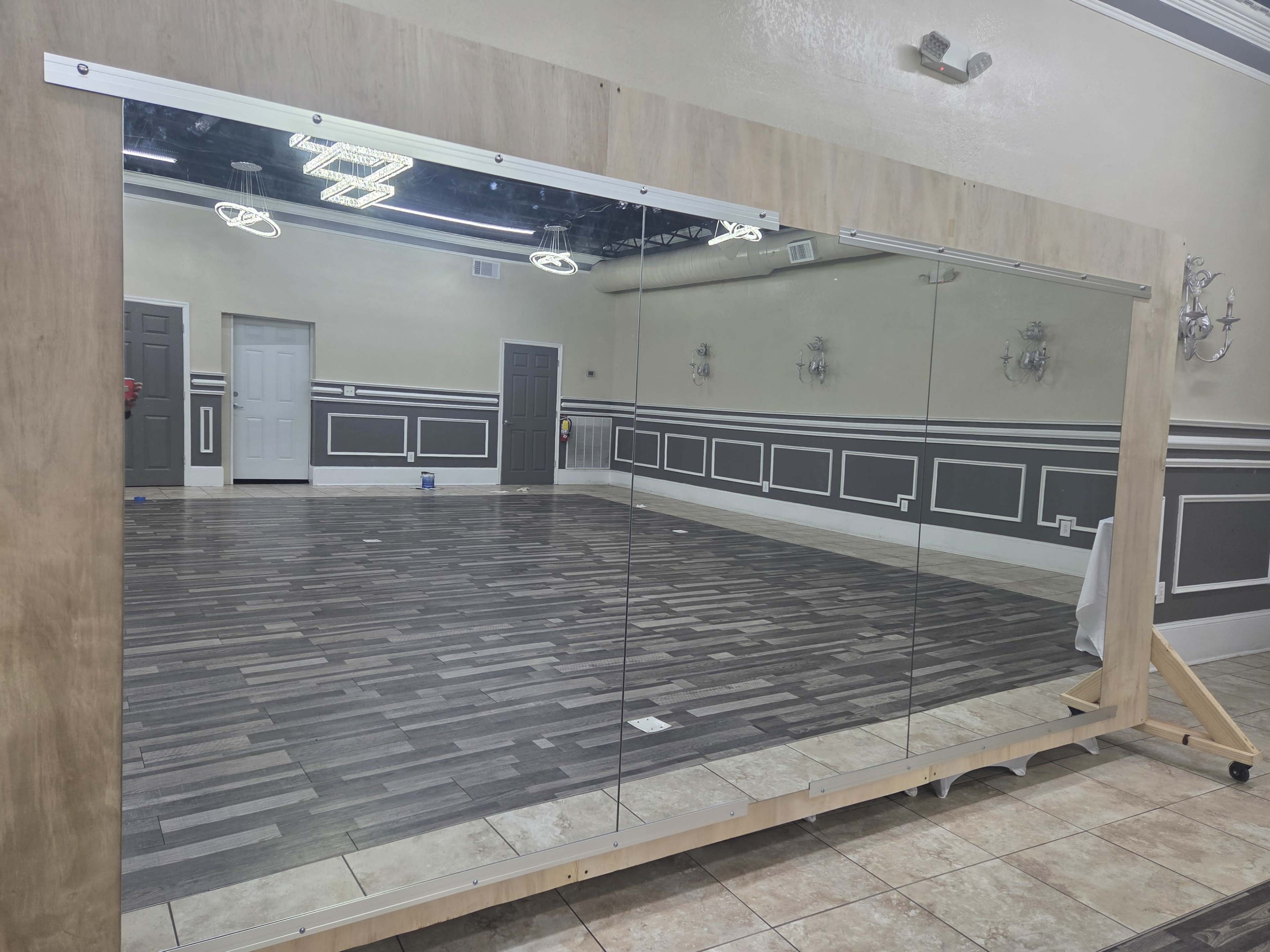 The image shows a large mirror mounted on a wooden frame against a wall in a spacious room with a tiled floor and decorative wall paneling.