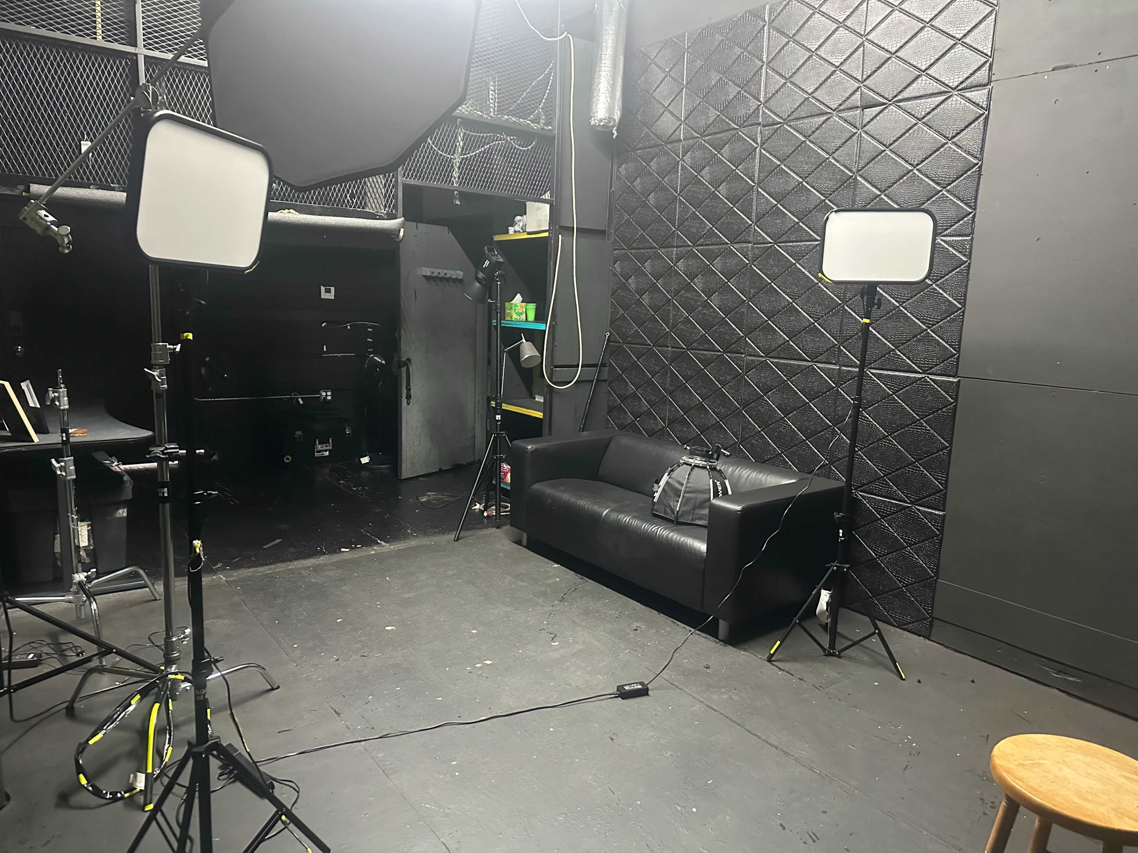 The image shows a dimly lit studio space with a black couch, two lighting stands with softboxes, and equipment against a textured black wall.