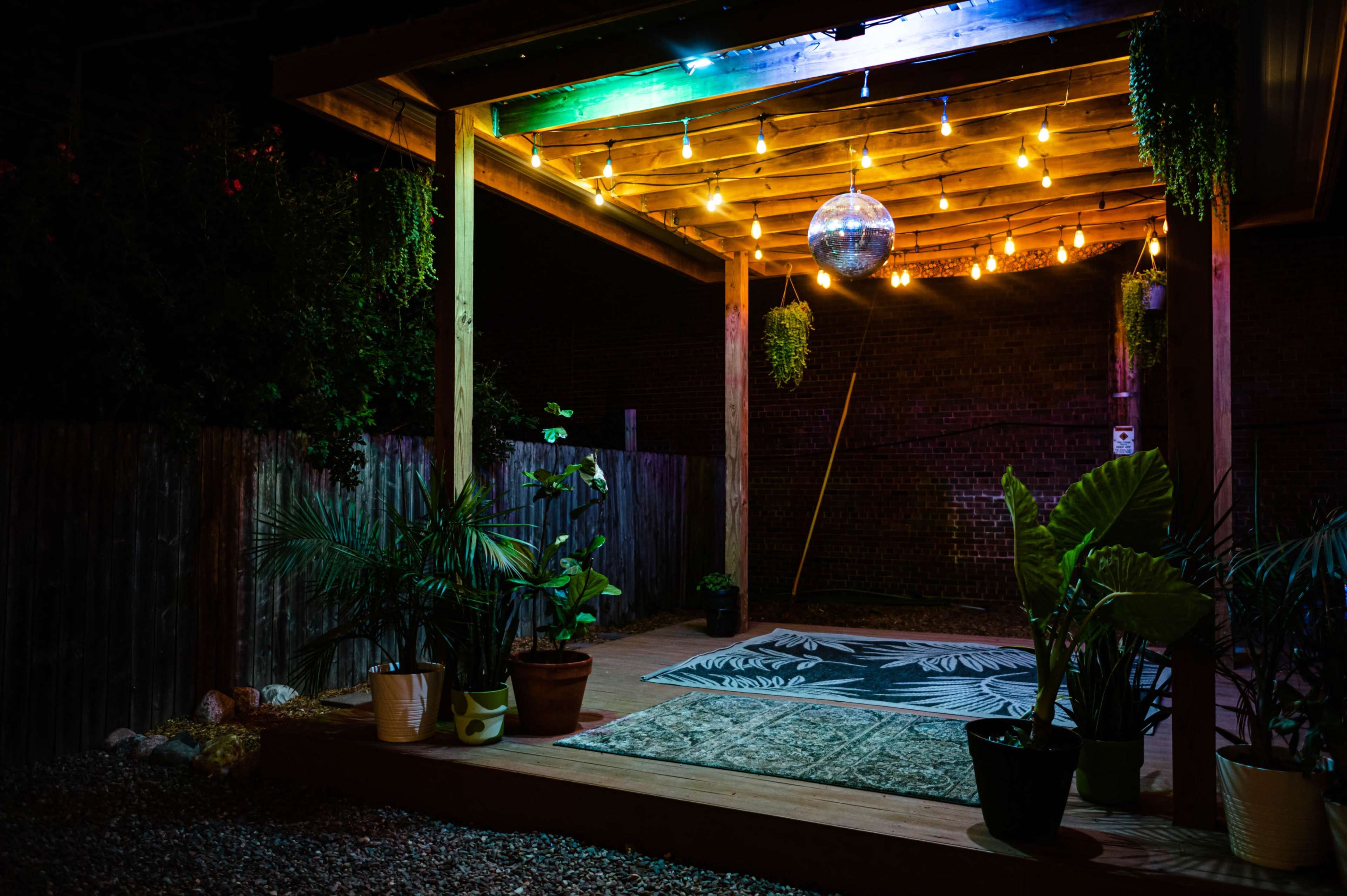 A wooden patio is illuminated by string lights and a disco ball, surrounded by potted plants and a patterned rug.