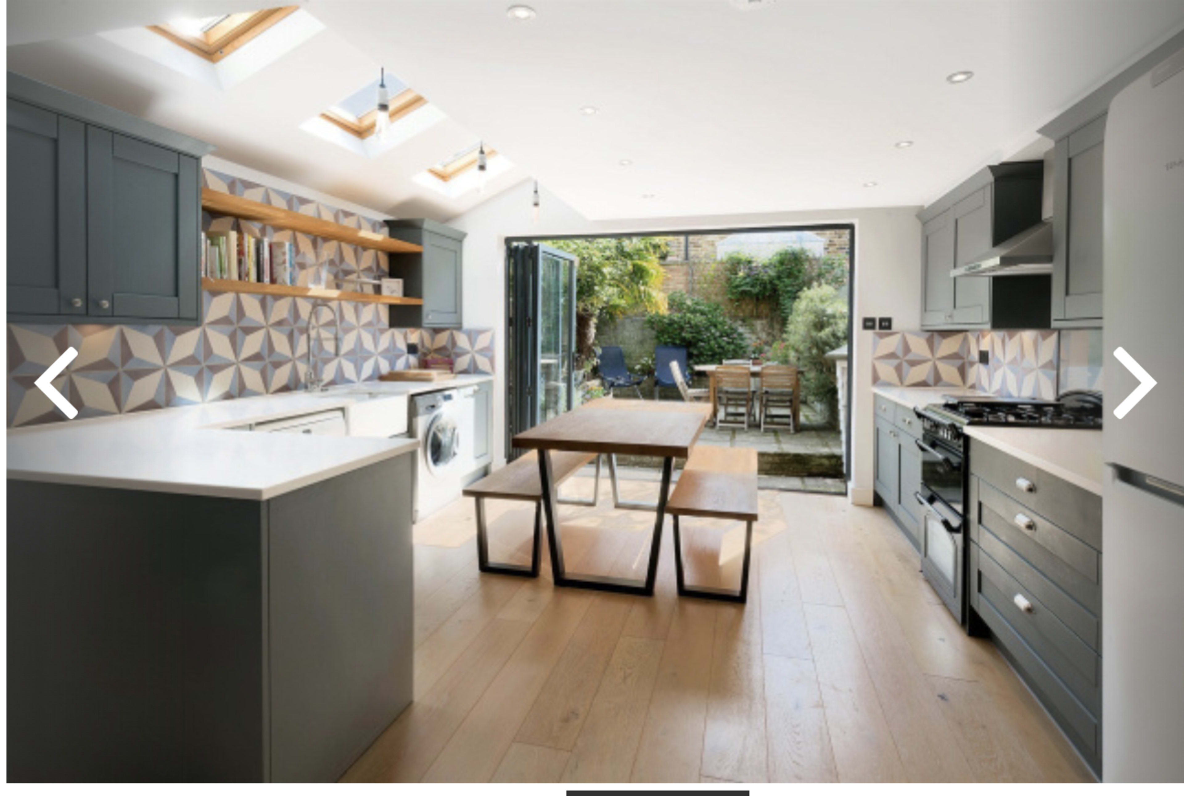 A modern kitchen features gray cabinetry, a central wooden dining table, and large glass doors opening to a garden.