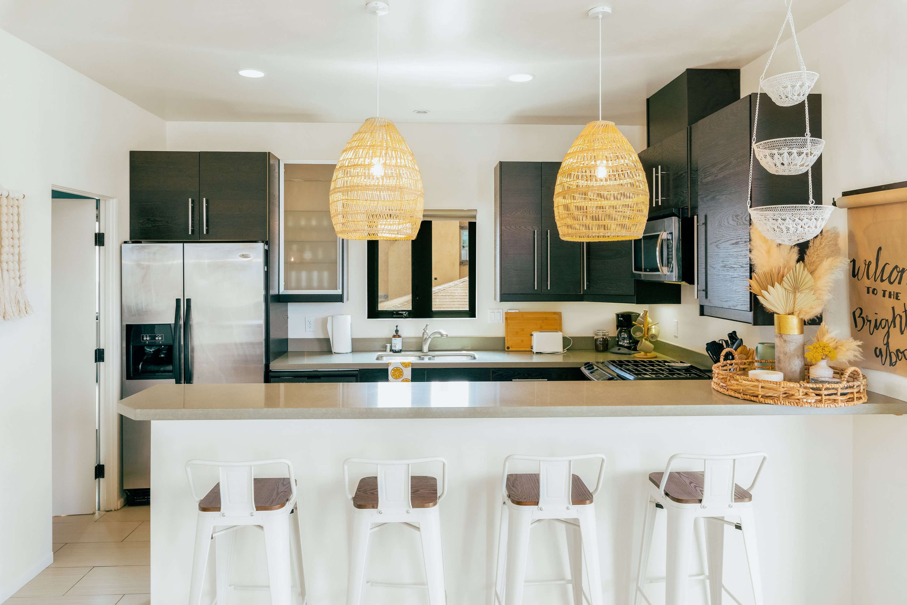 A modern kitchen with a breakfast bar, featuring wooden pendant lights, dark cabinetry, stainless steel appliances, and a white countertop.
