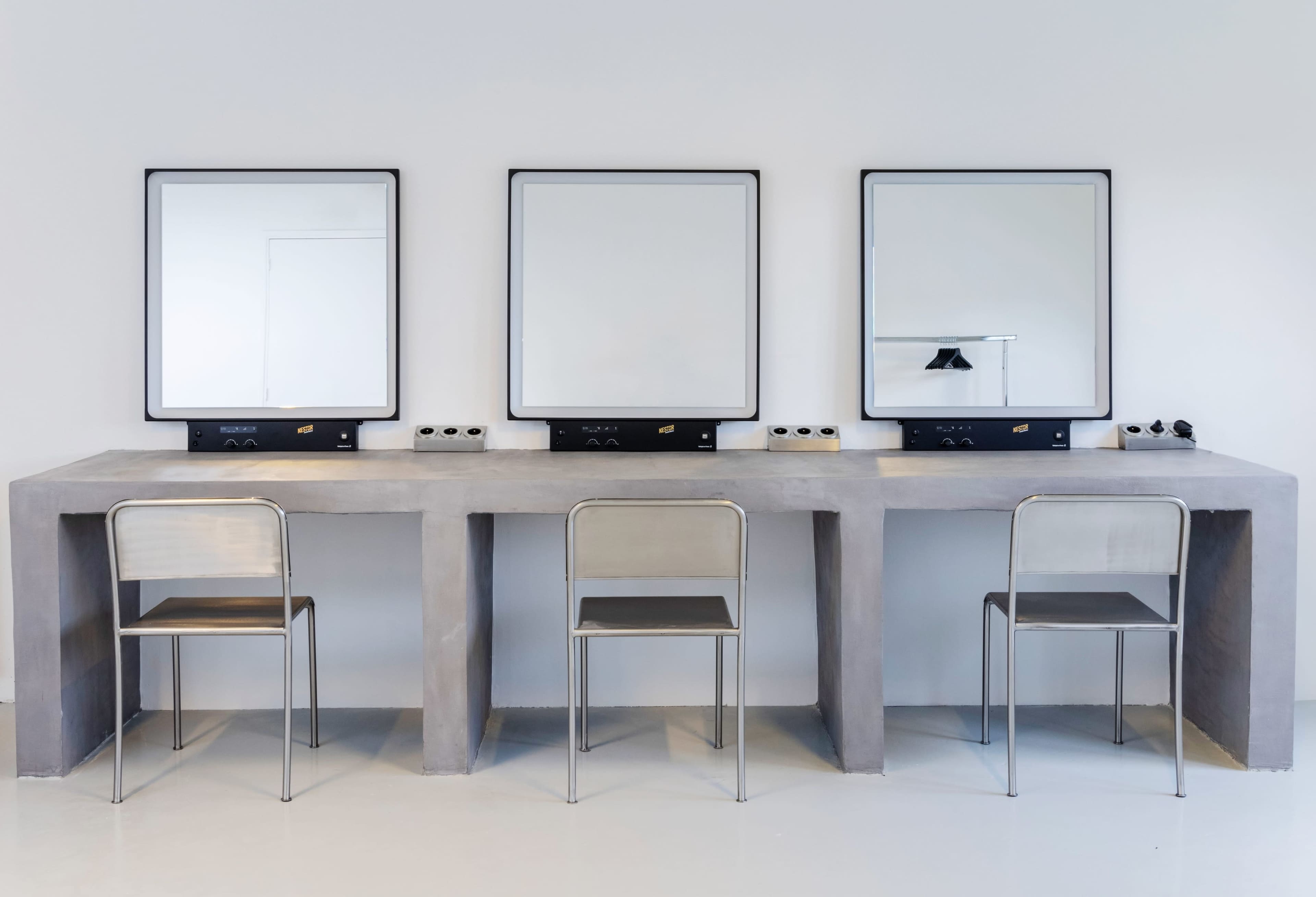A minimalist vanity area with three mirrors above a concrete countertop and three metal chairs.