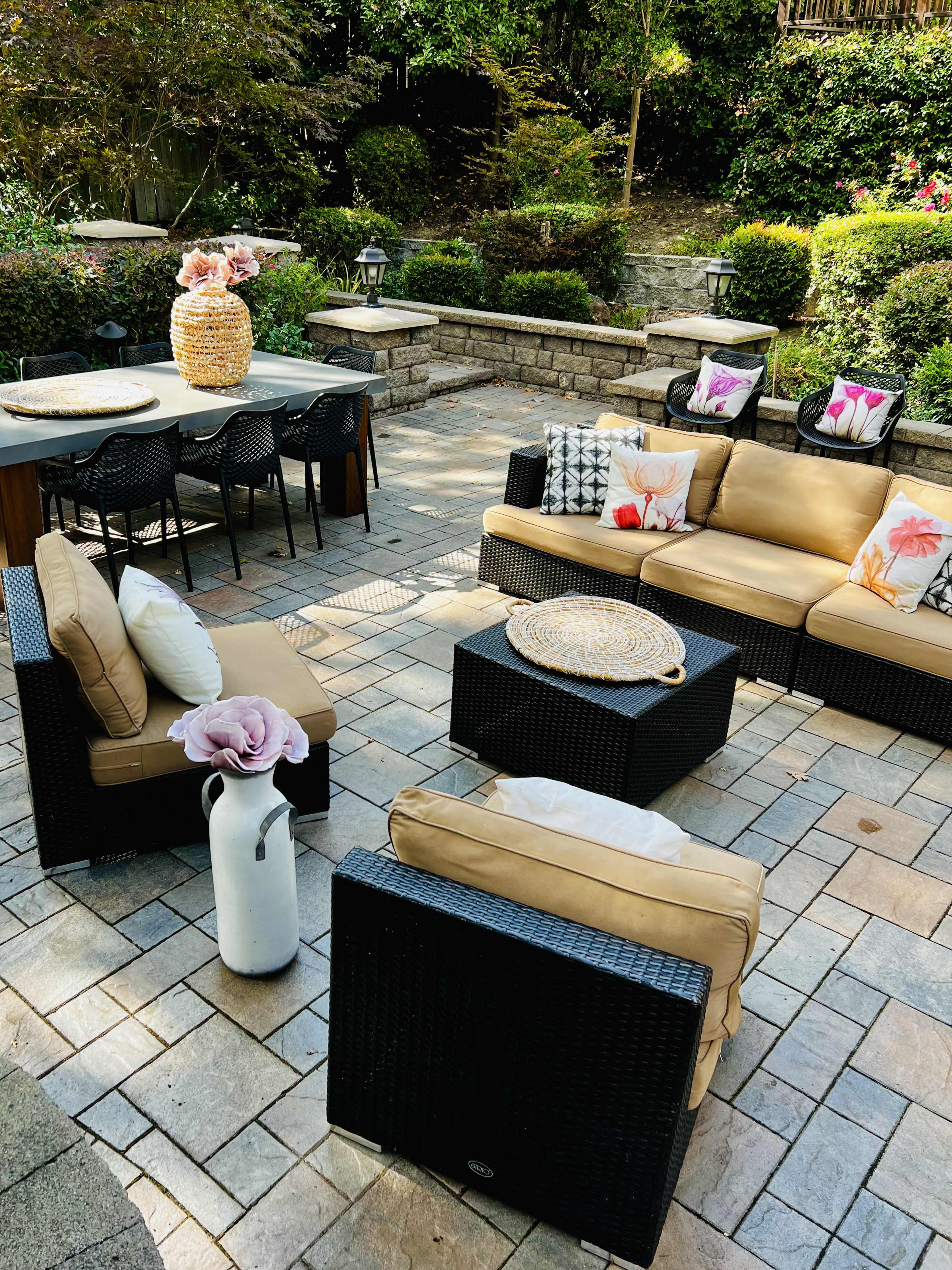 A patio area features a seating arrangement with light-colored sofas and black wicker chairs, a coffee table, and a dining table set amidst landscaped greenery.