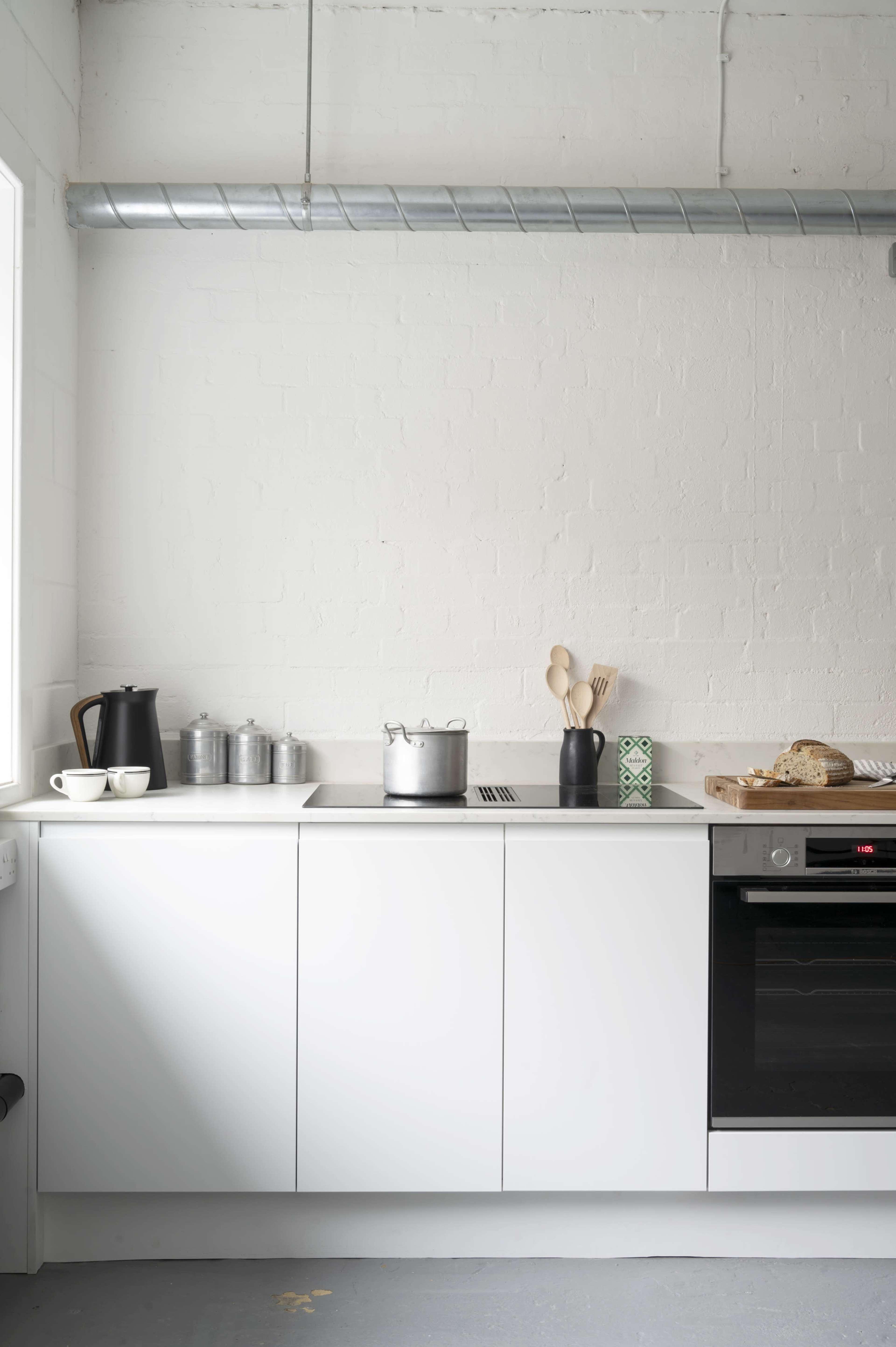 A modern kitchen features a white countertop with a pot on a stovetop, a black kettle, metal canisters, wooden utensils, and a loaf of bread on a tray.