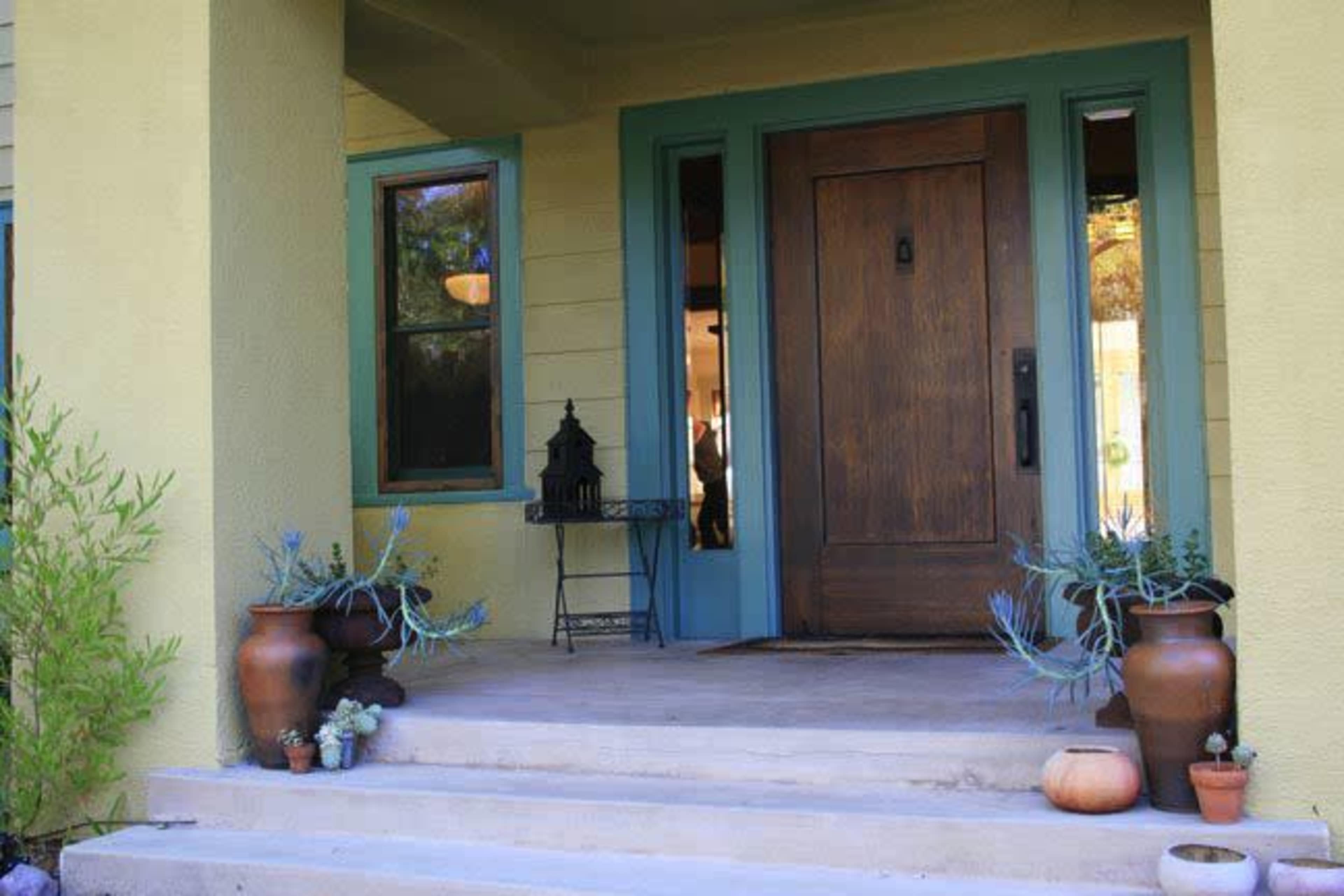 A front porch with a wooden door, green trim, and potted plants on either side of the steps.