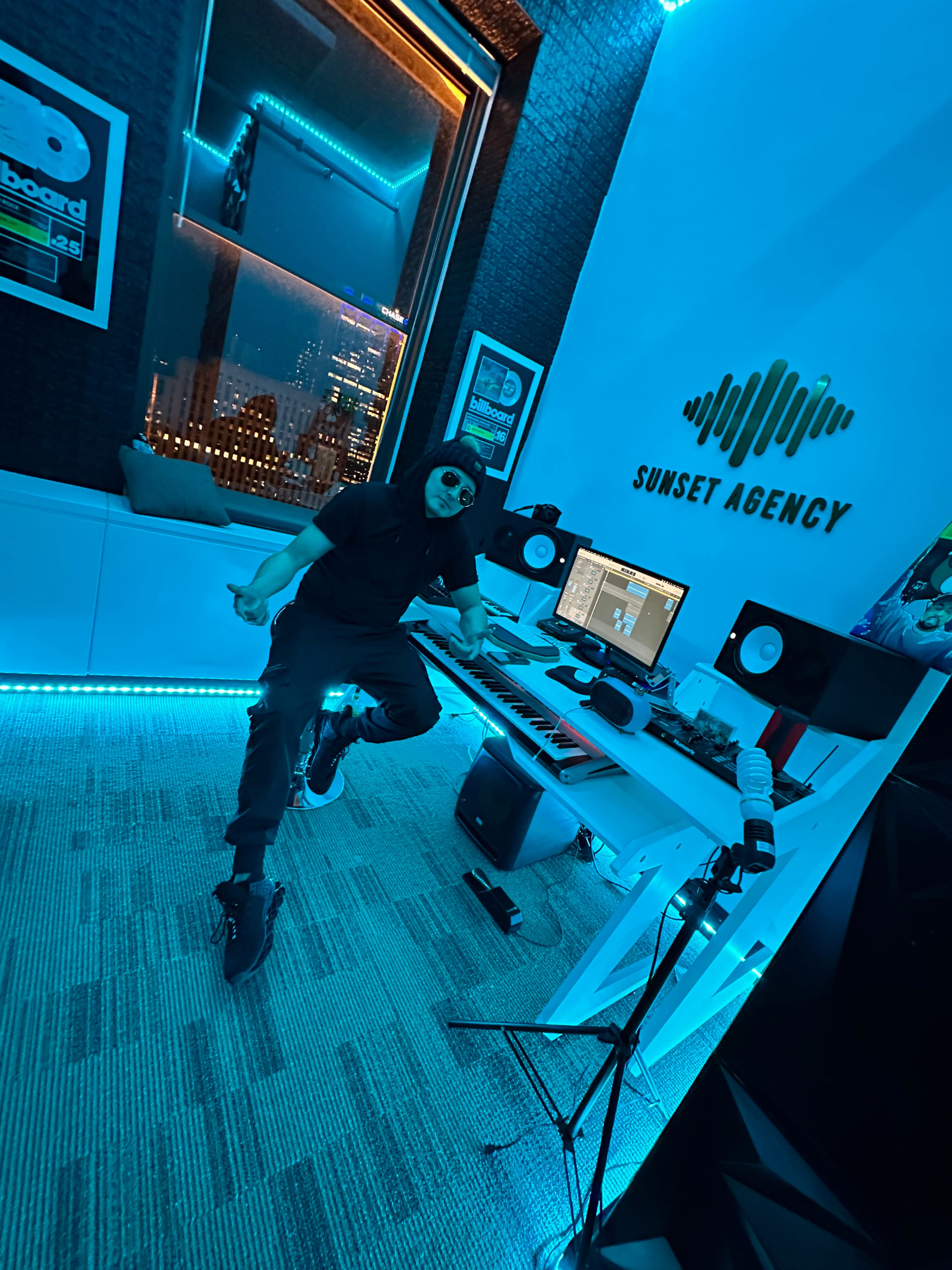 A person poses in a modern music production studio with blue ambient lighting, surrounded by equipment and a computer setup.