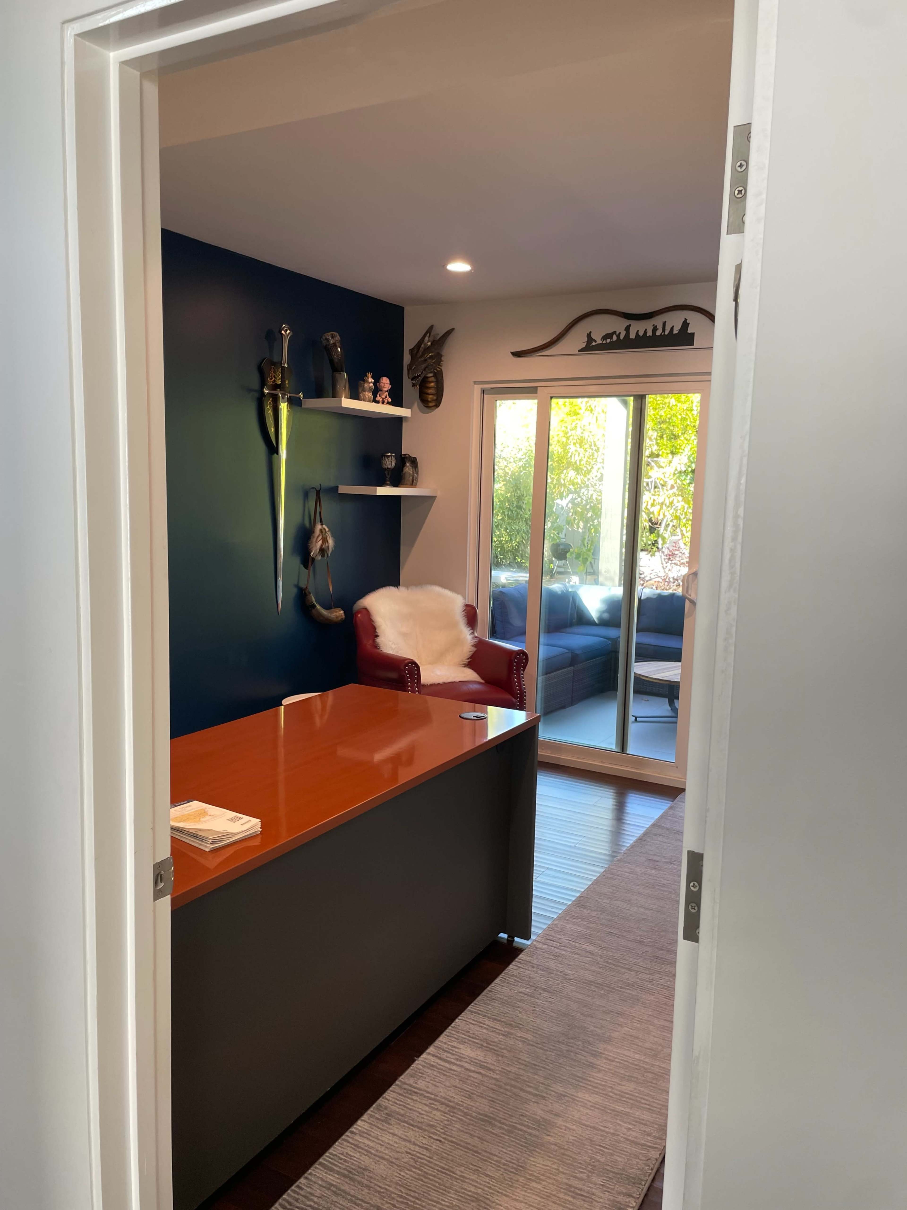 The image shows an office space with a wooden desk, a chair in the corner, and sliding glass doors leading to an outdoor area.