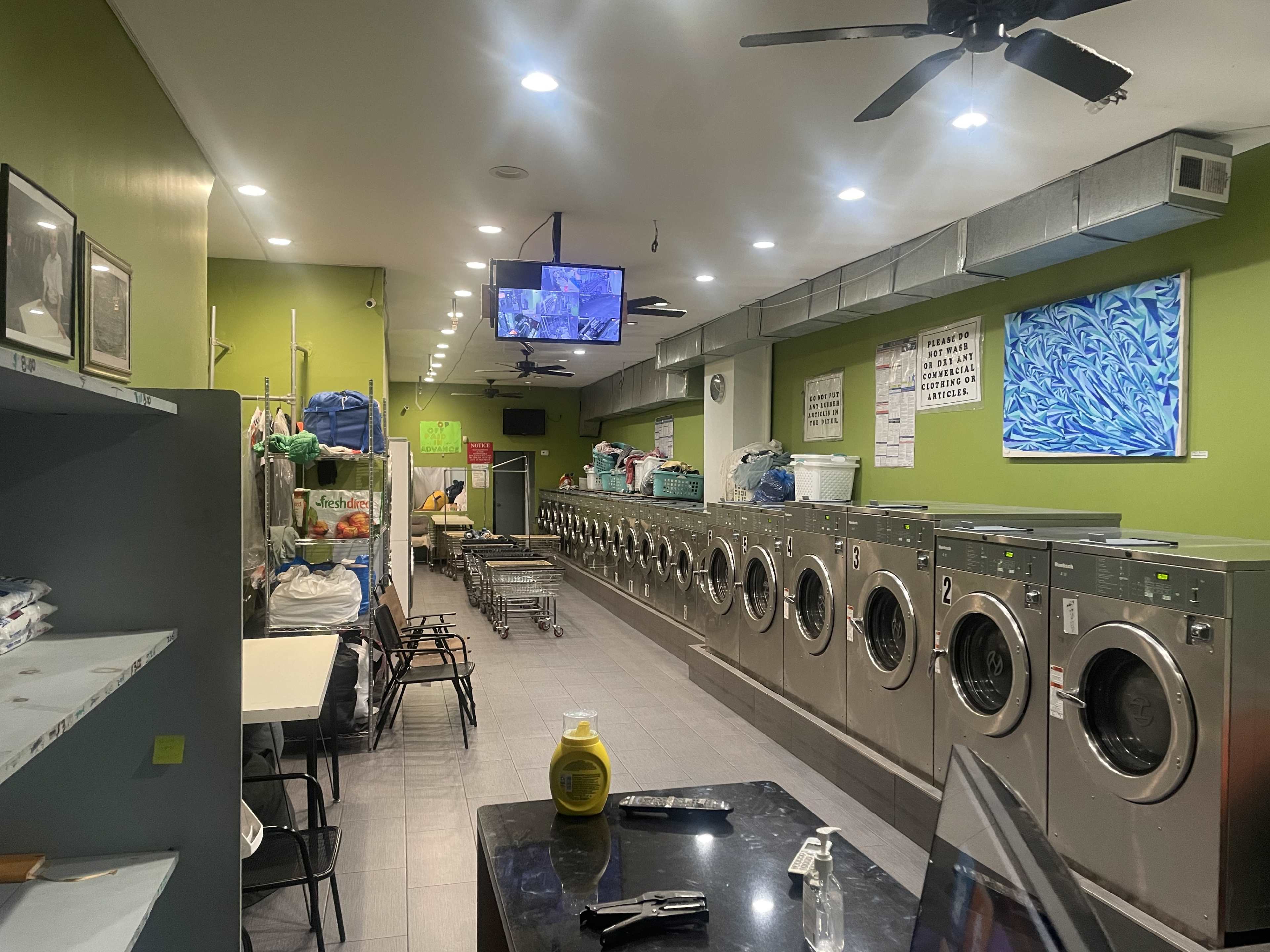 A brightly lit laundromat features multiple washing machines along one side, with folding tables and chairs on the opposite side, and a television mounted on the wall.