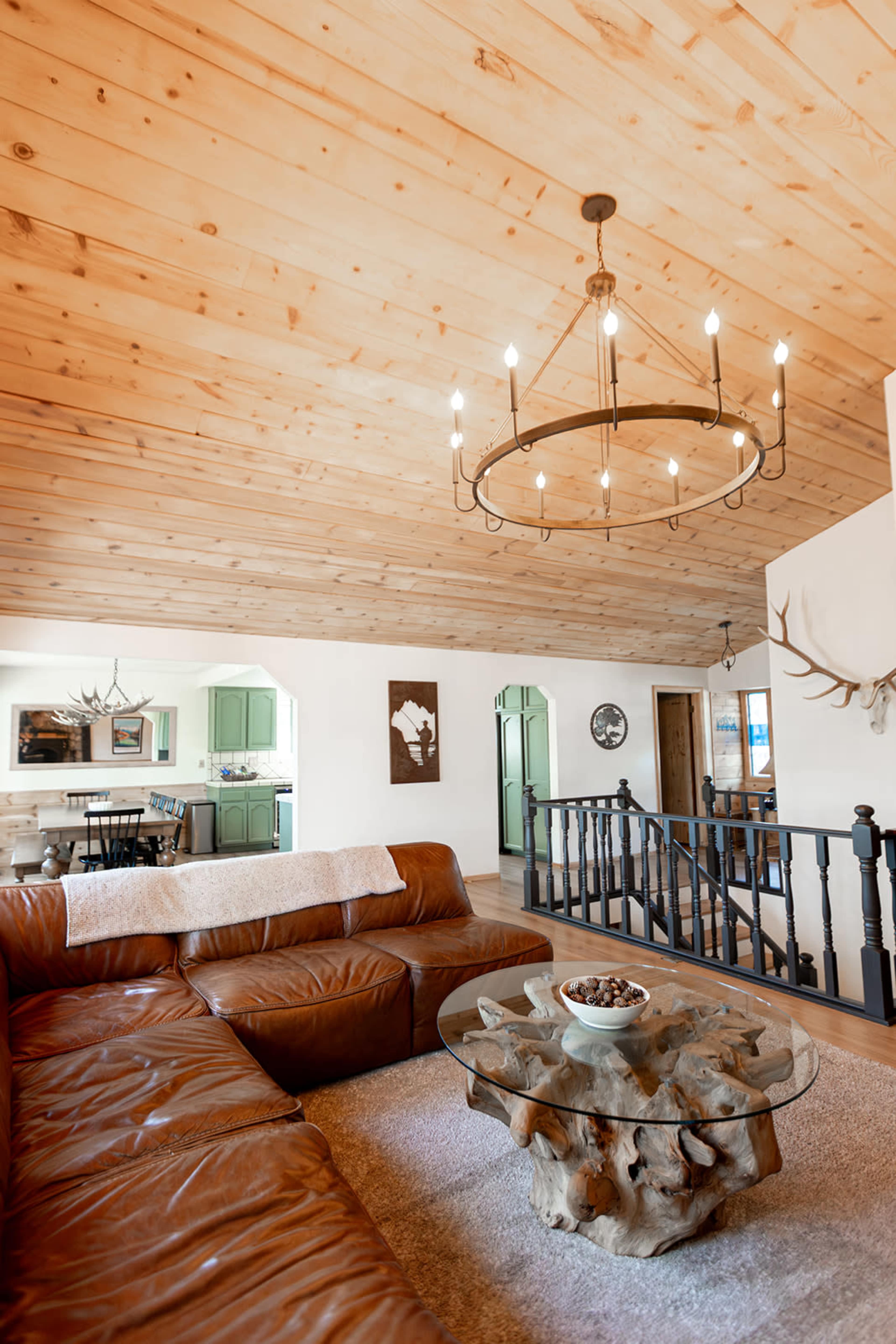 The image shows a spacious living room with wooden ceilings, a leather sectional sofa, a circular chandelier, and a unique coffee table made of wood.