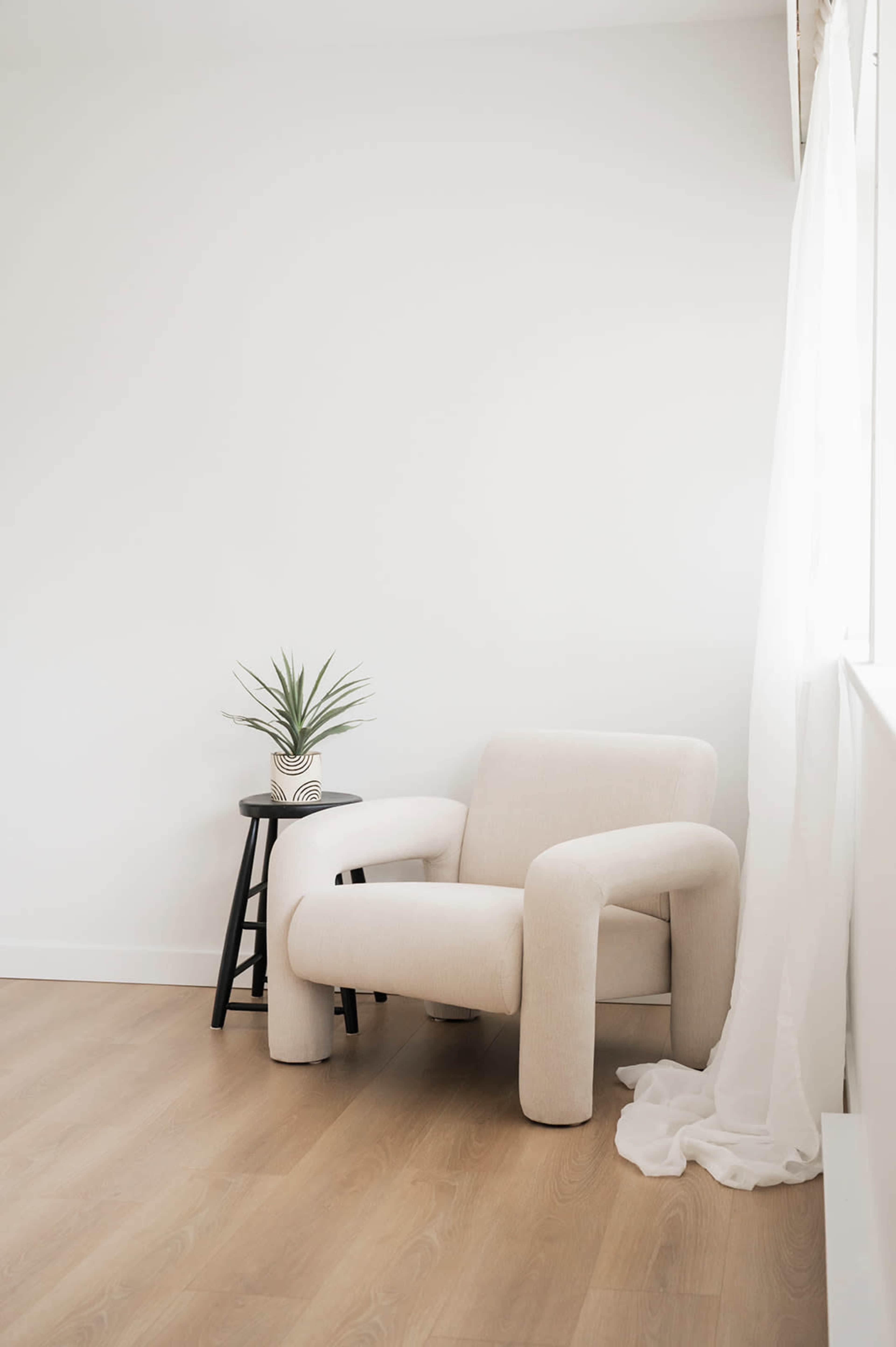 A beige, armless chair is positioned next to a black stool with a potted plant on top, against a light-colored wall and wooden floor.