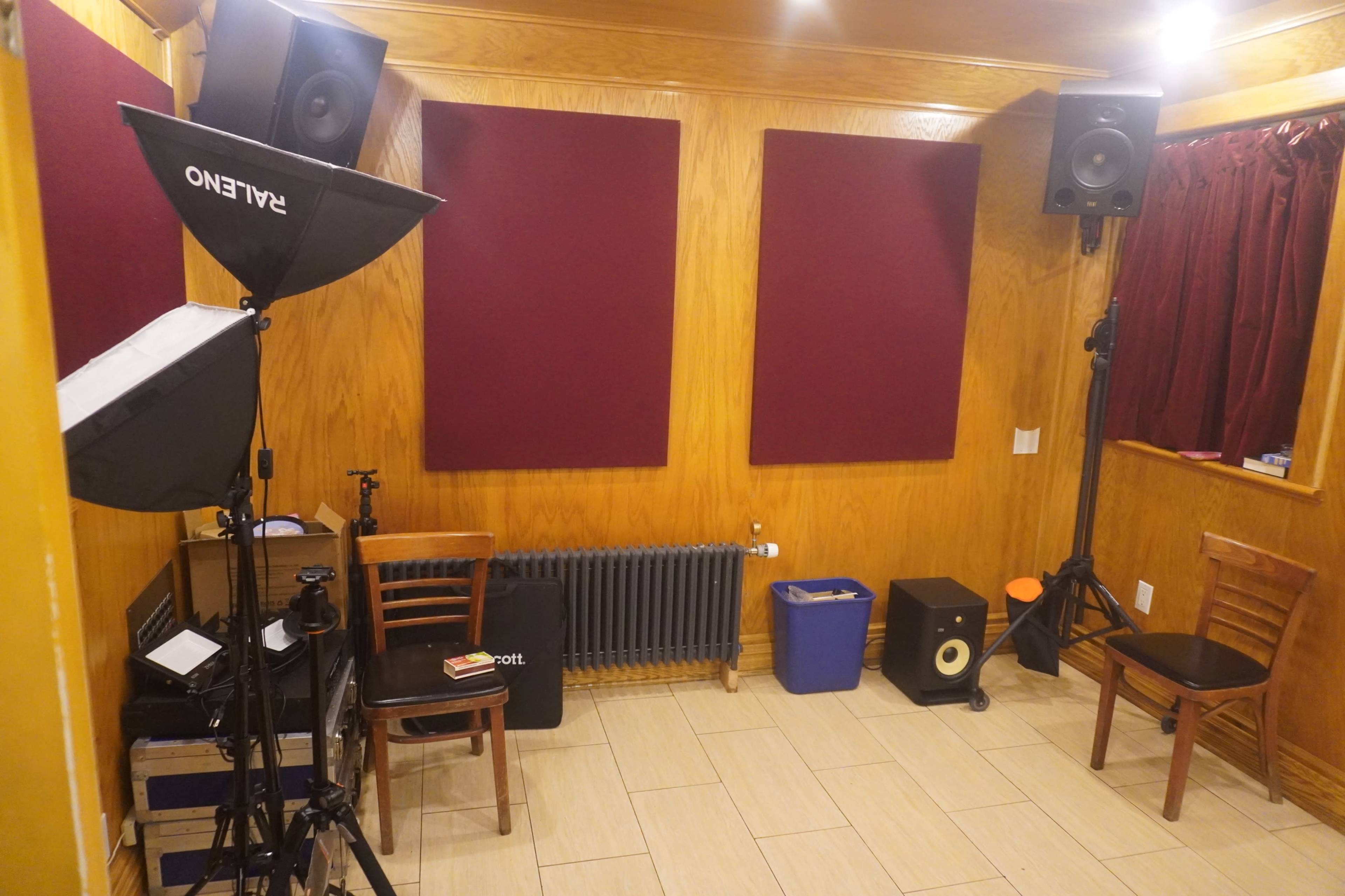 A small, soundproofed recording studio with acoustic panels on the walls, microphones on stands, and speakers positioned in the corners, along with chairs and equipment.