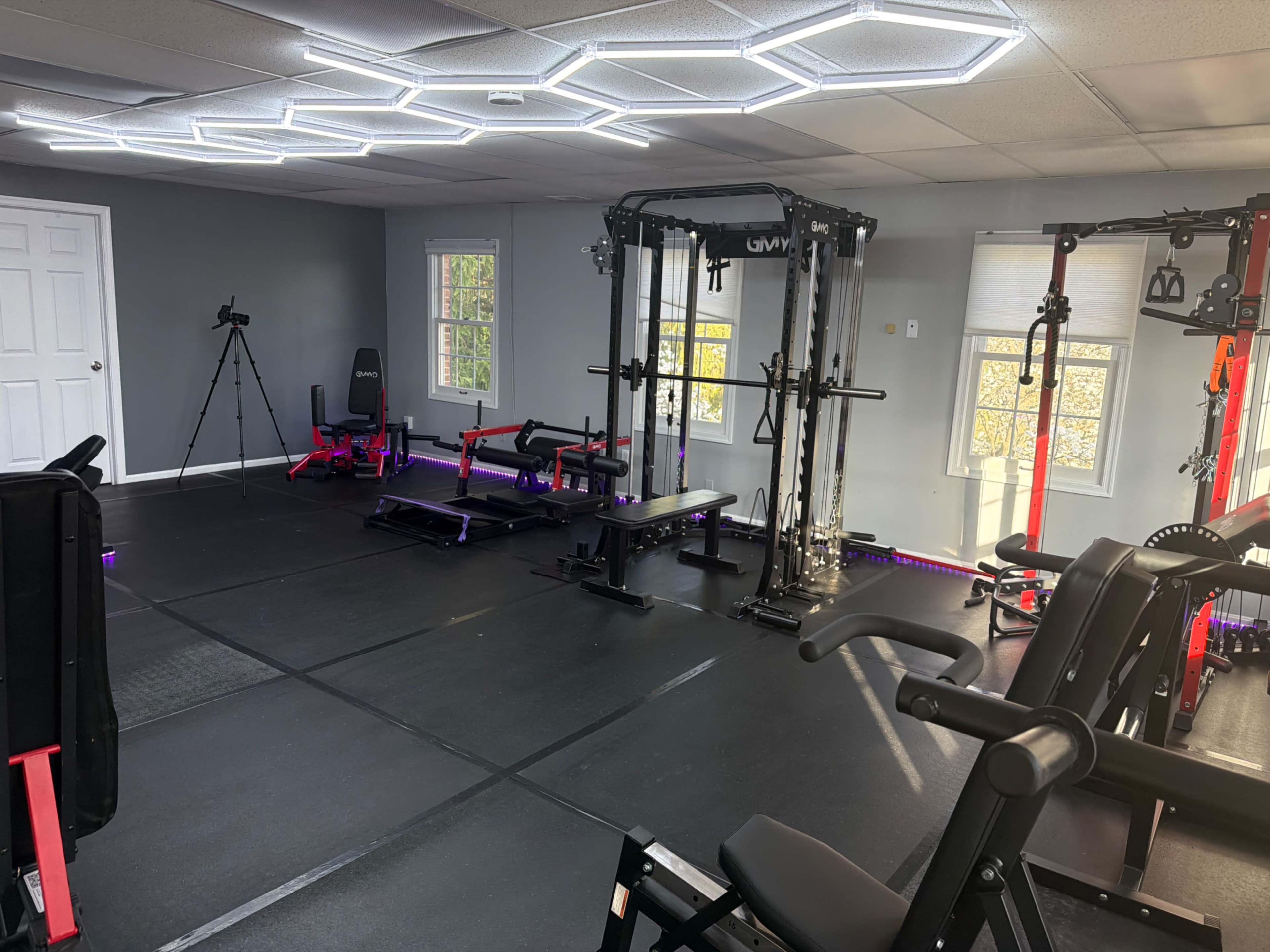 The image shows a well-equipped home gym featuring various exercise machines, benches, and weights, along with a tripod for a camera.
