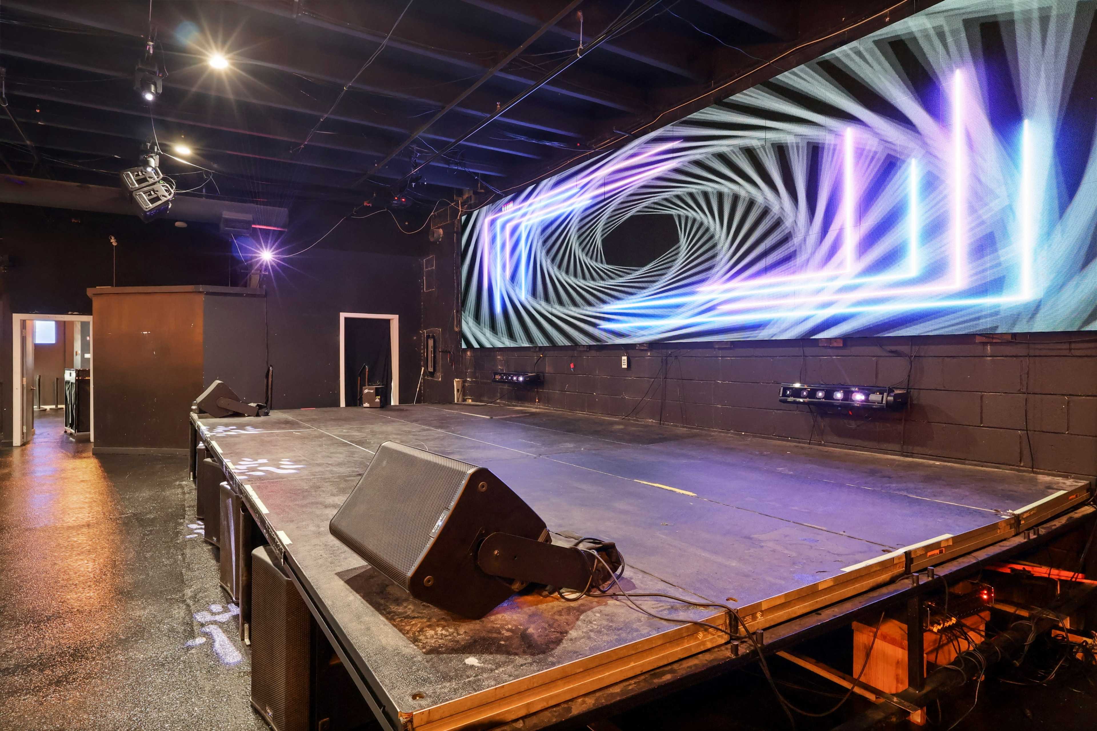 A stage is set up with a large screens displaying dynamic visuals, surrounded by dark walls and minimal lighting.