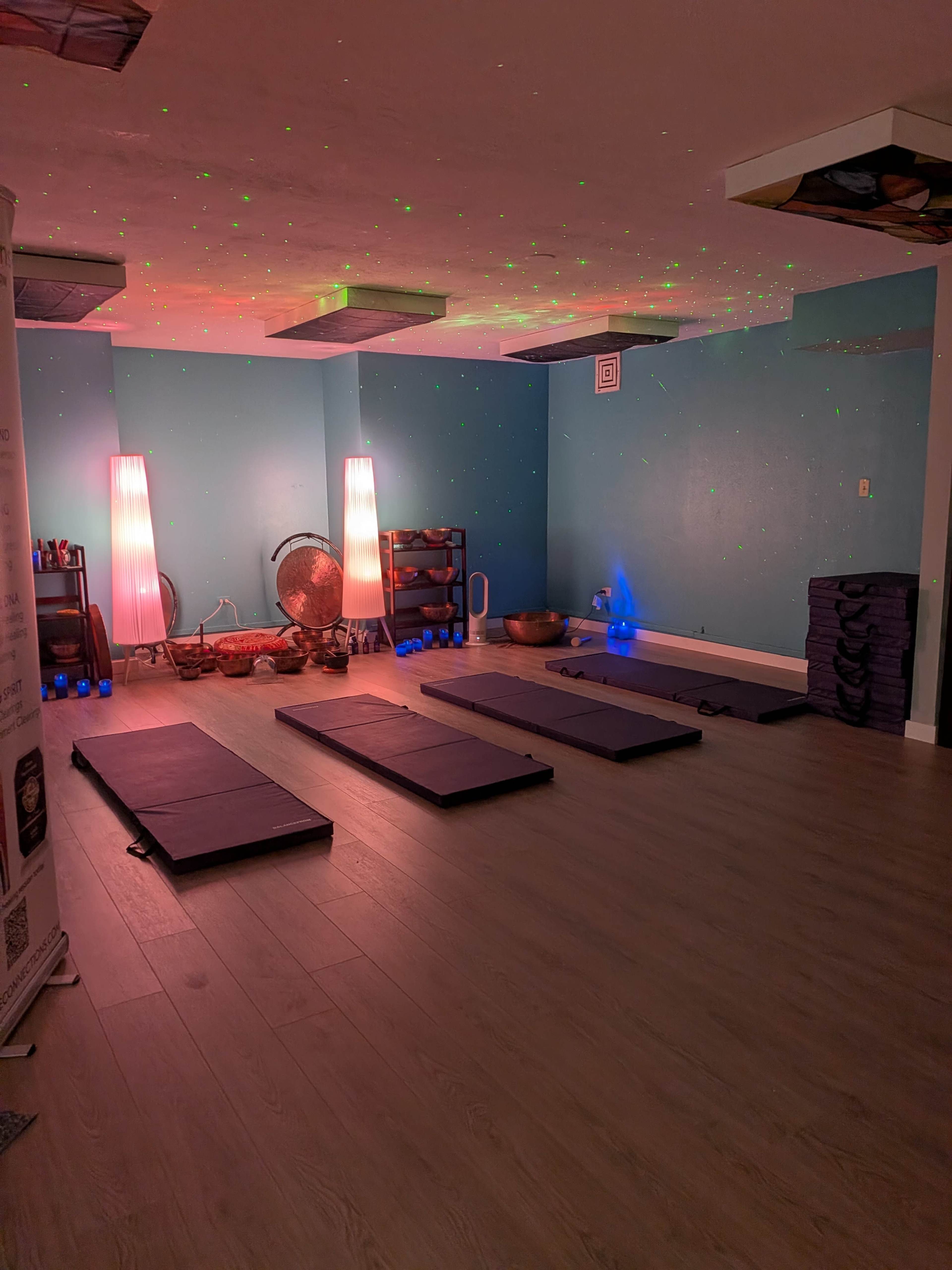 The image shows a dimly lit yoga studio with exercise mats arranged on the floor, illuminated by colored lights and accompanied by various wellness props.