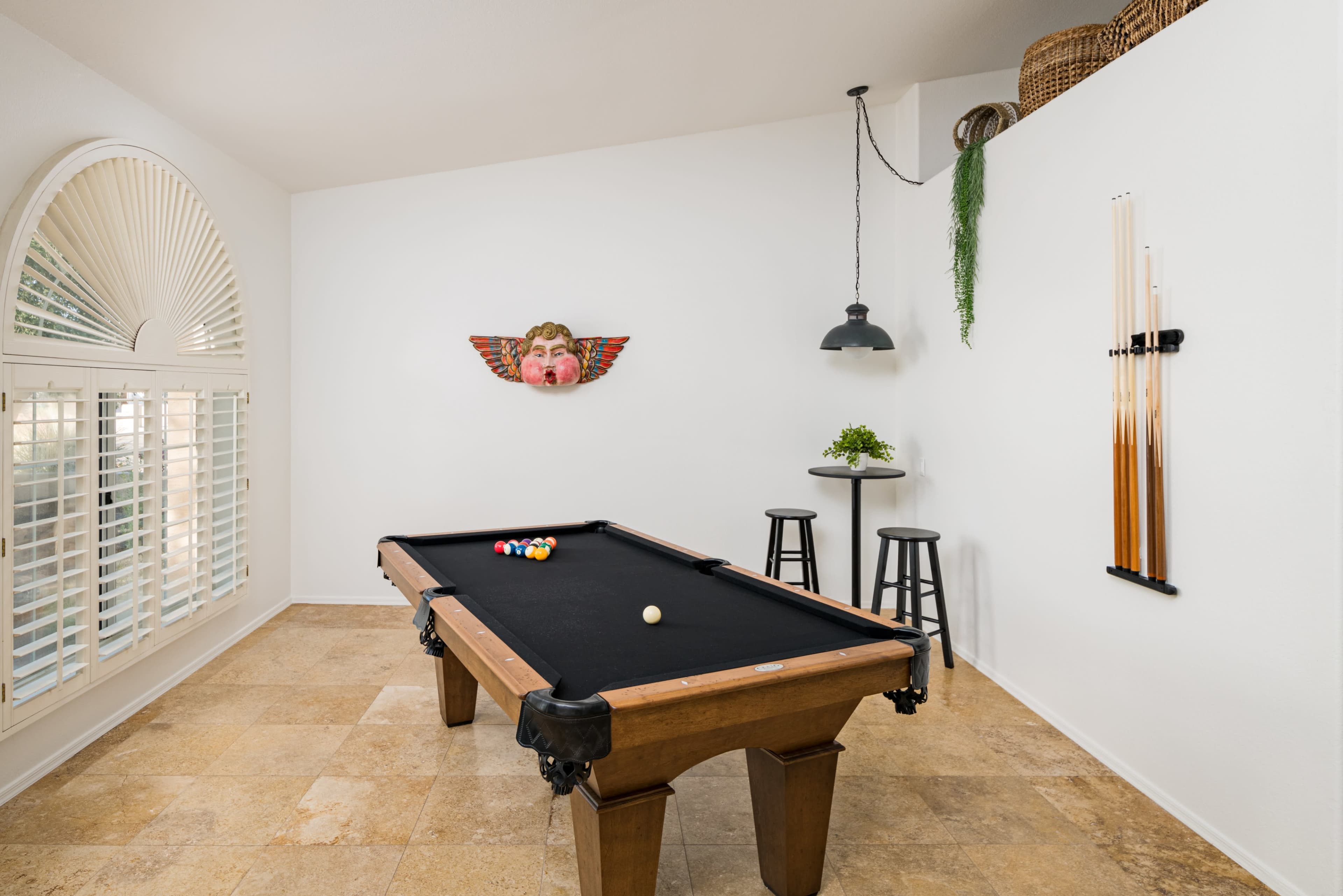 The image shows a room with a billiard table, two bar stools, and wall decor, including a face mask, set against white walls and a window with shutters.