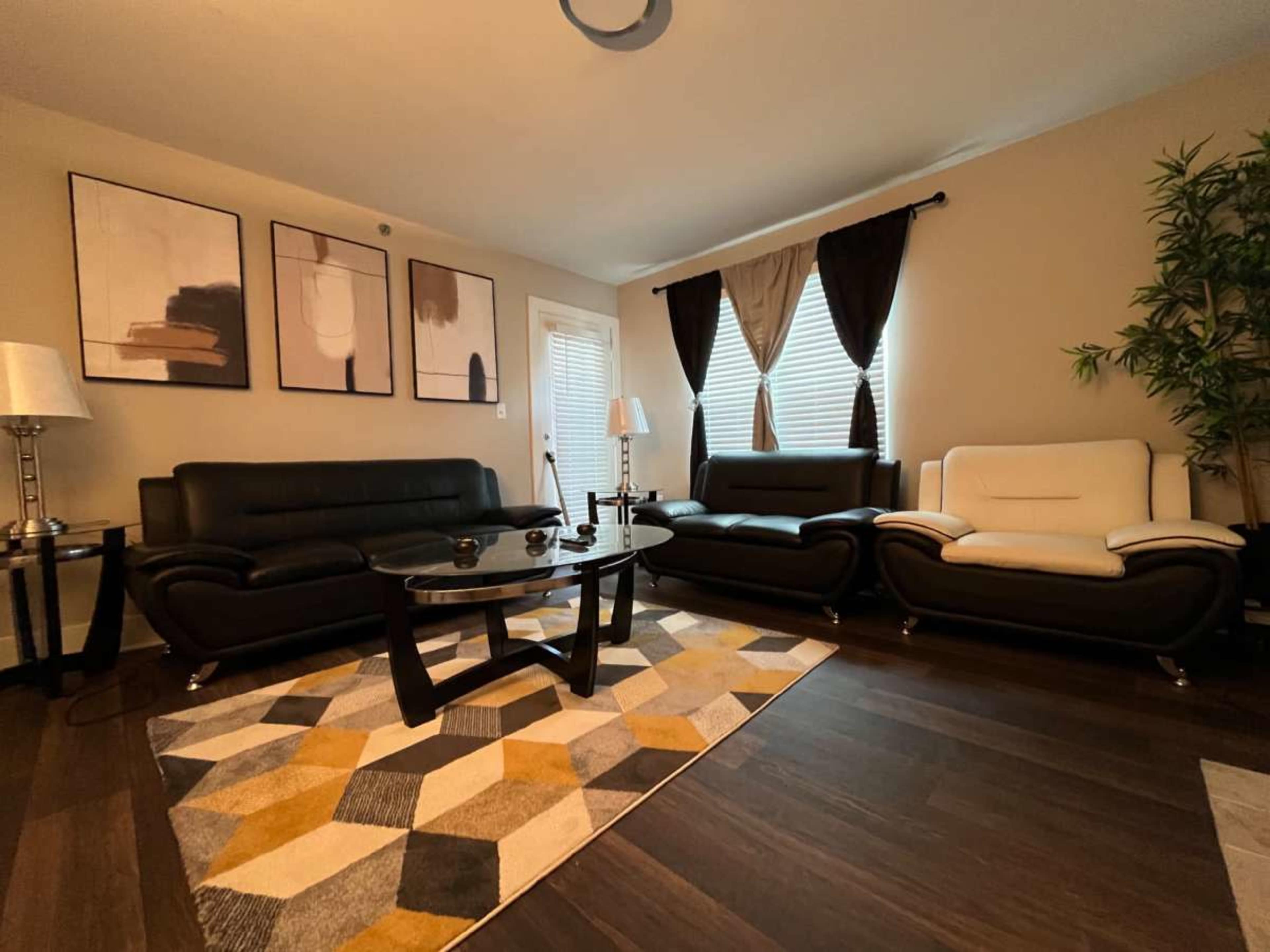 The image shows a modern living room with three sofas, a glass coffee table, and framed wall art, illuminated by natural light.