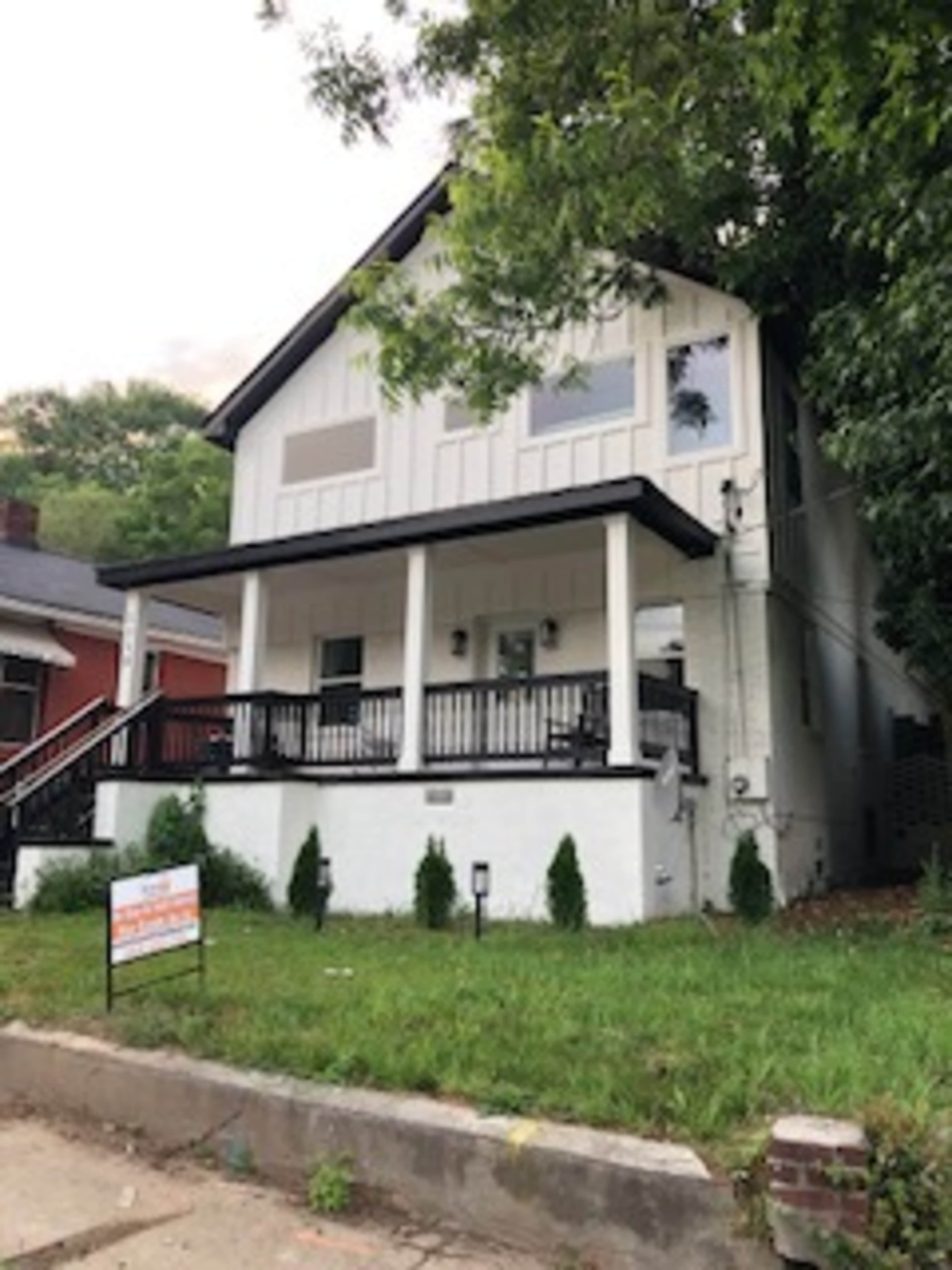 A two-story white house with a porch and black railing is for sale, surrounded by green grass and small bushes.