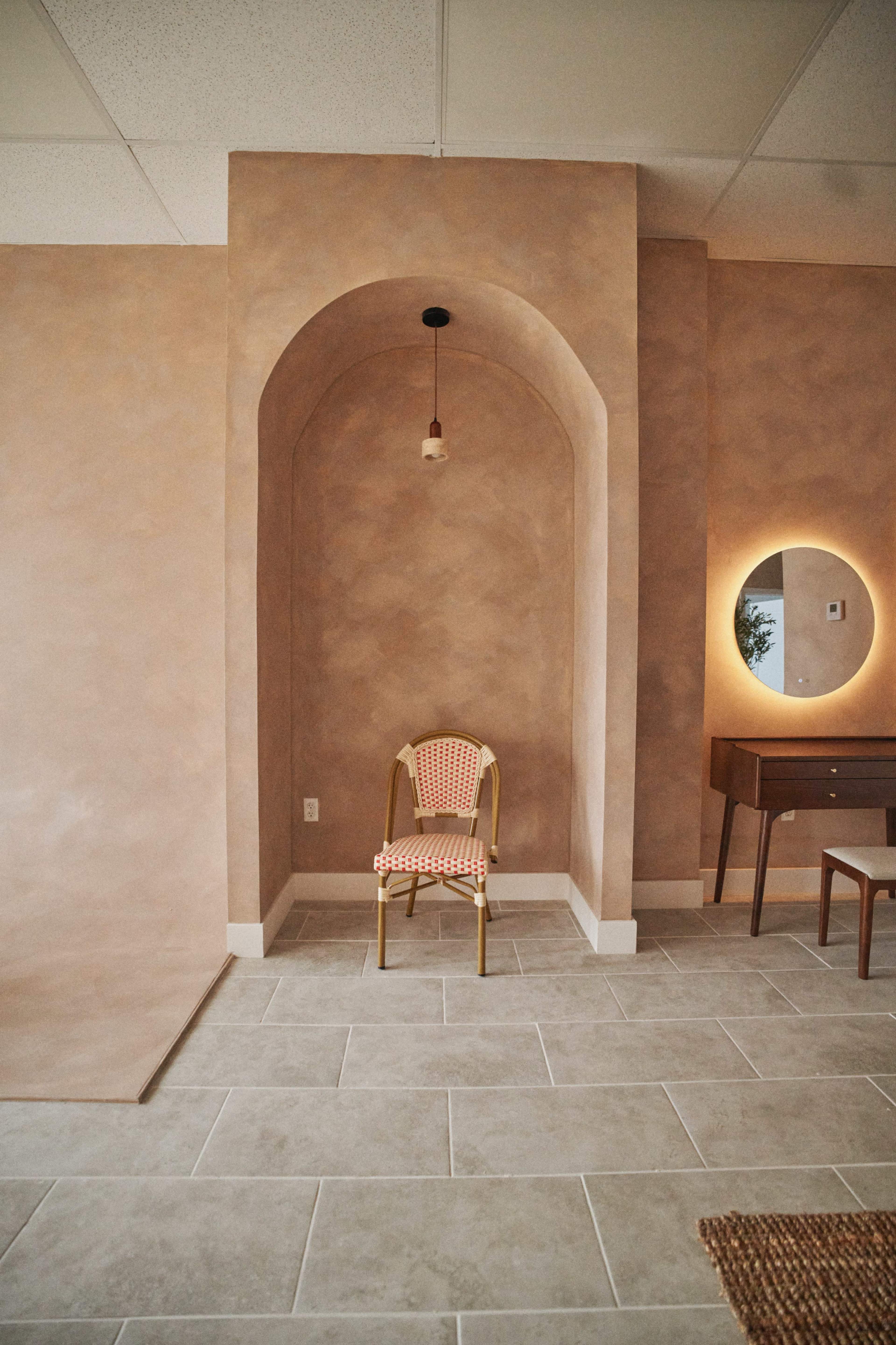 The image shows a minimalist interior space featuring a chair with a checkered pattern placed in a textured alcove, complemented by a round mirror and a wooden table.