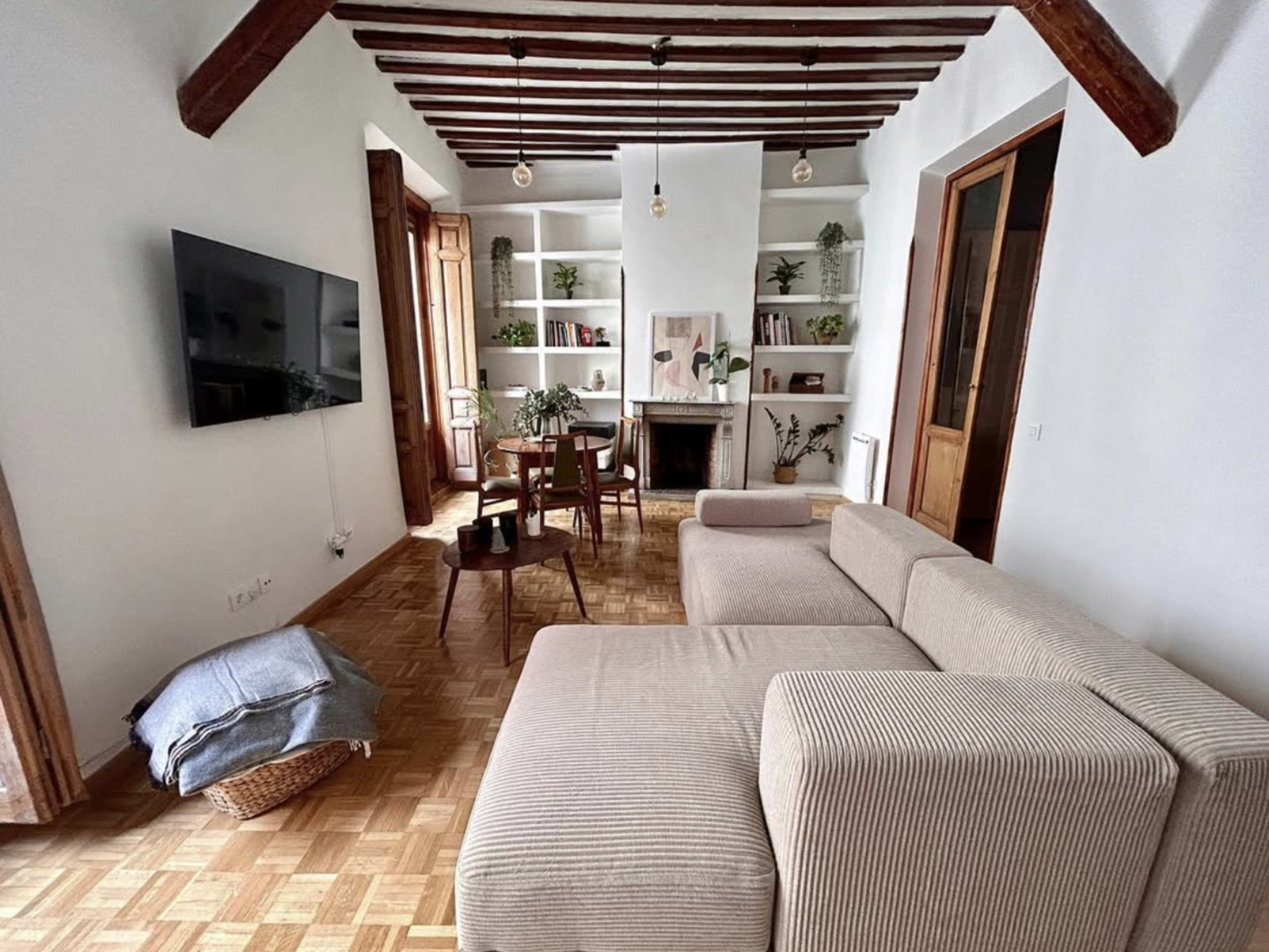 The image shows a living room with a sectional sofa, a television mounted on the wall, a small dining table, and shelves with plants and decorations.