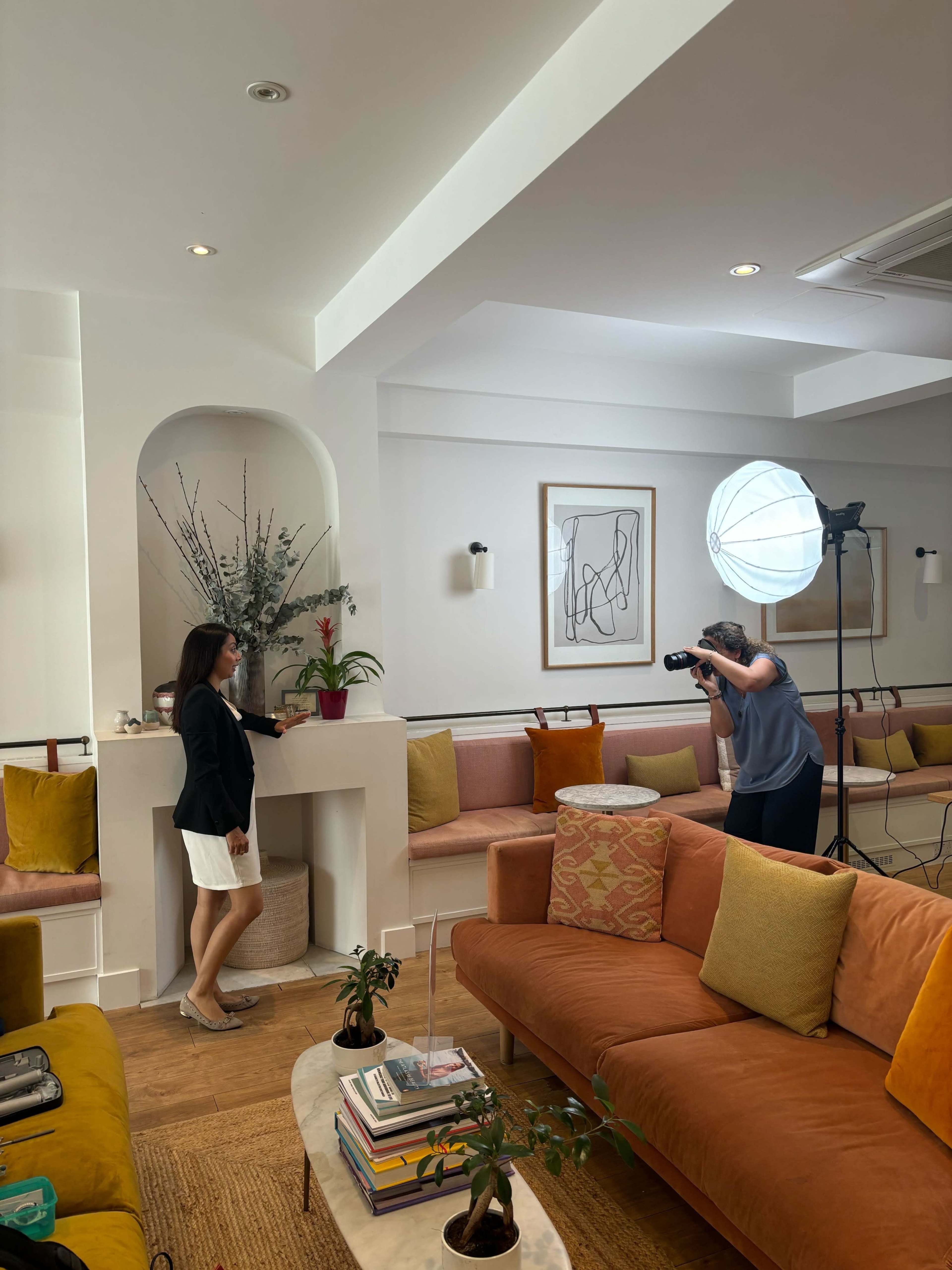 A woman poses by a decorative fireplace while another woman photographs her in a stylish, well-lit sitting area.