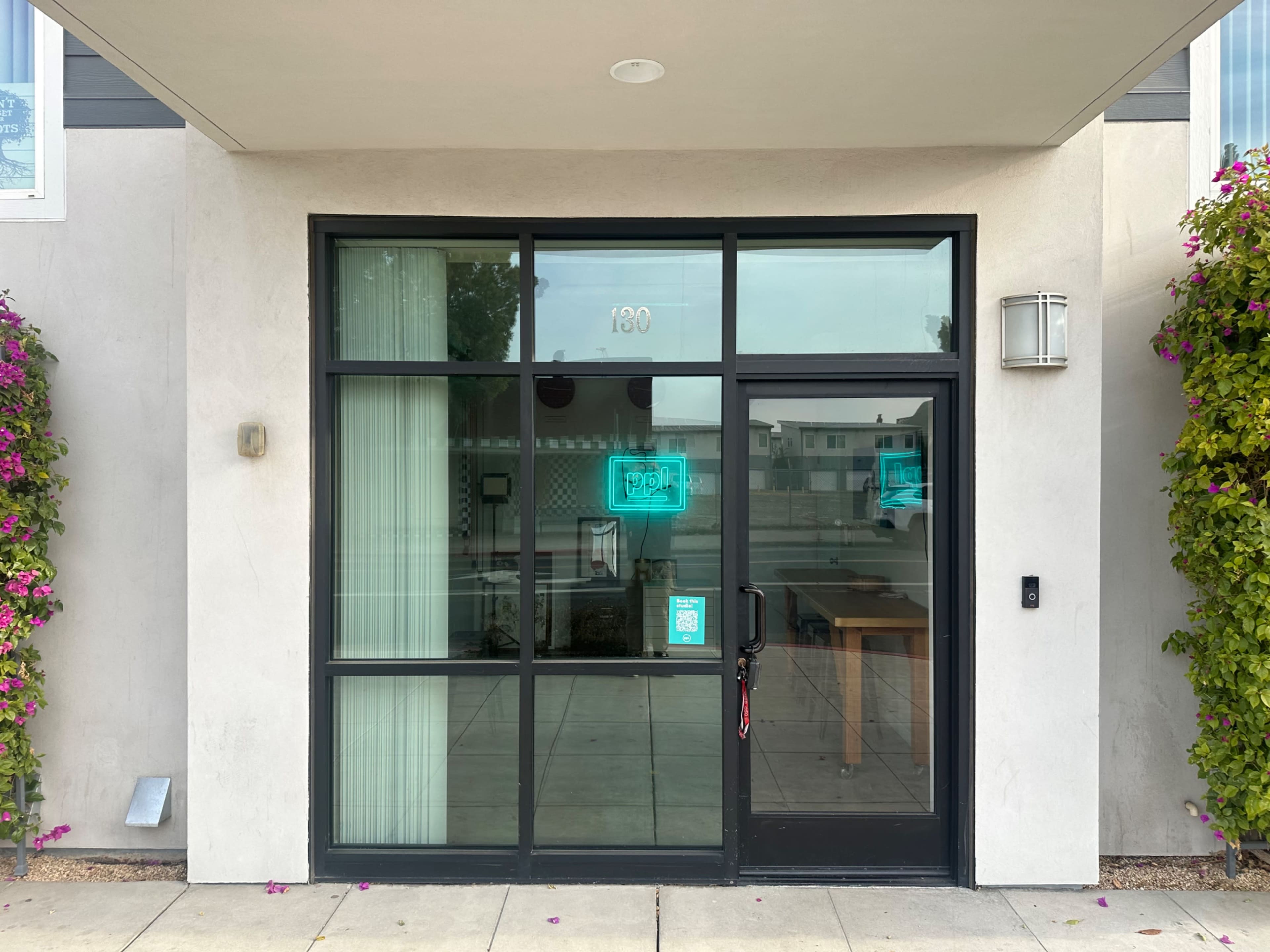 A modern entrance with large glass doors, a light blue neon sign, and flowering vines on either side of the entrance.