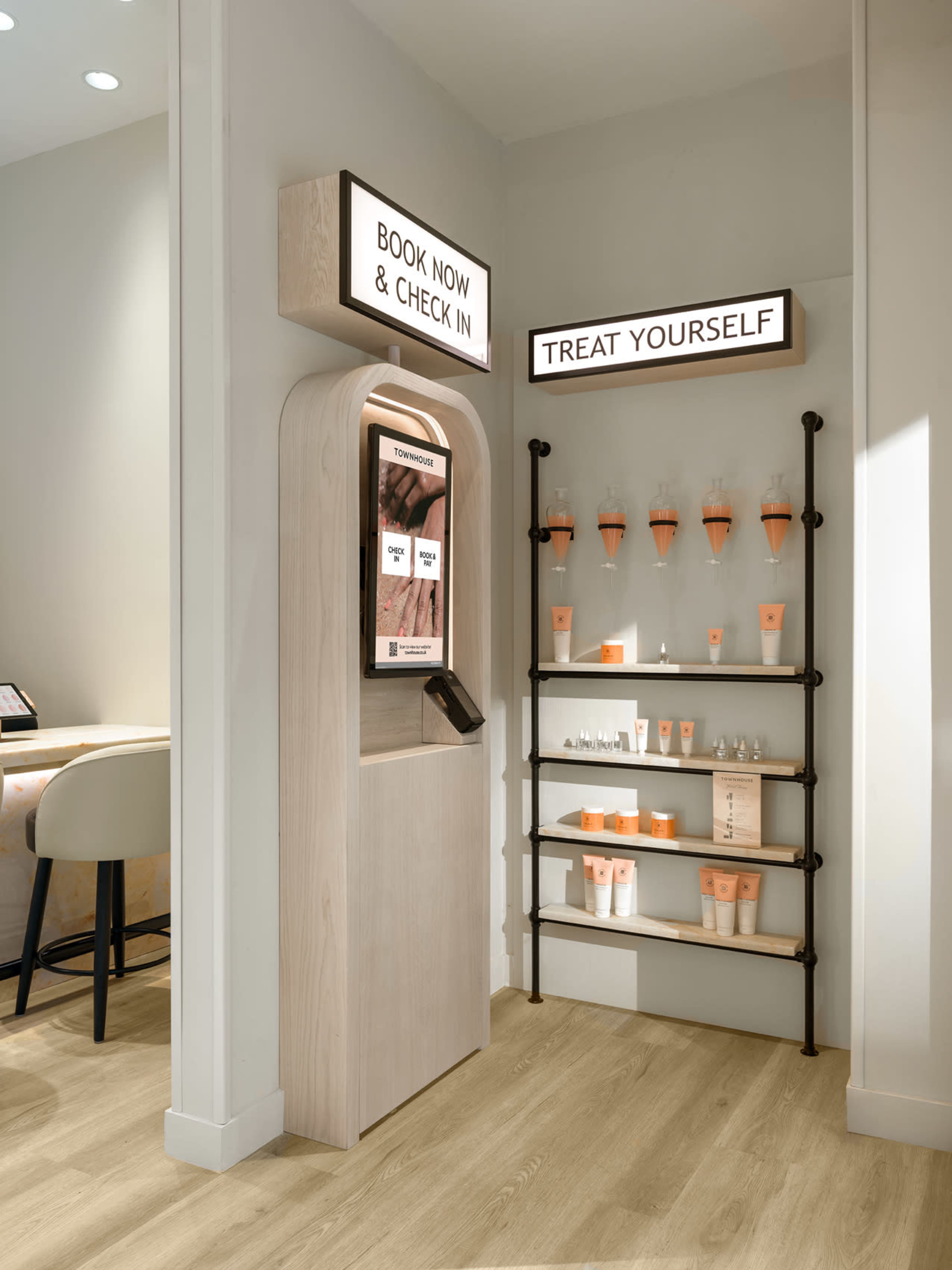 The image displays a modern reception area featuring a digital kiosk for booking and check-in, alongside a display shelf stocked with skincare products.
