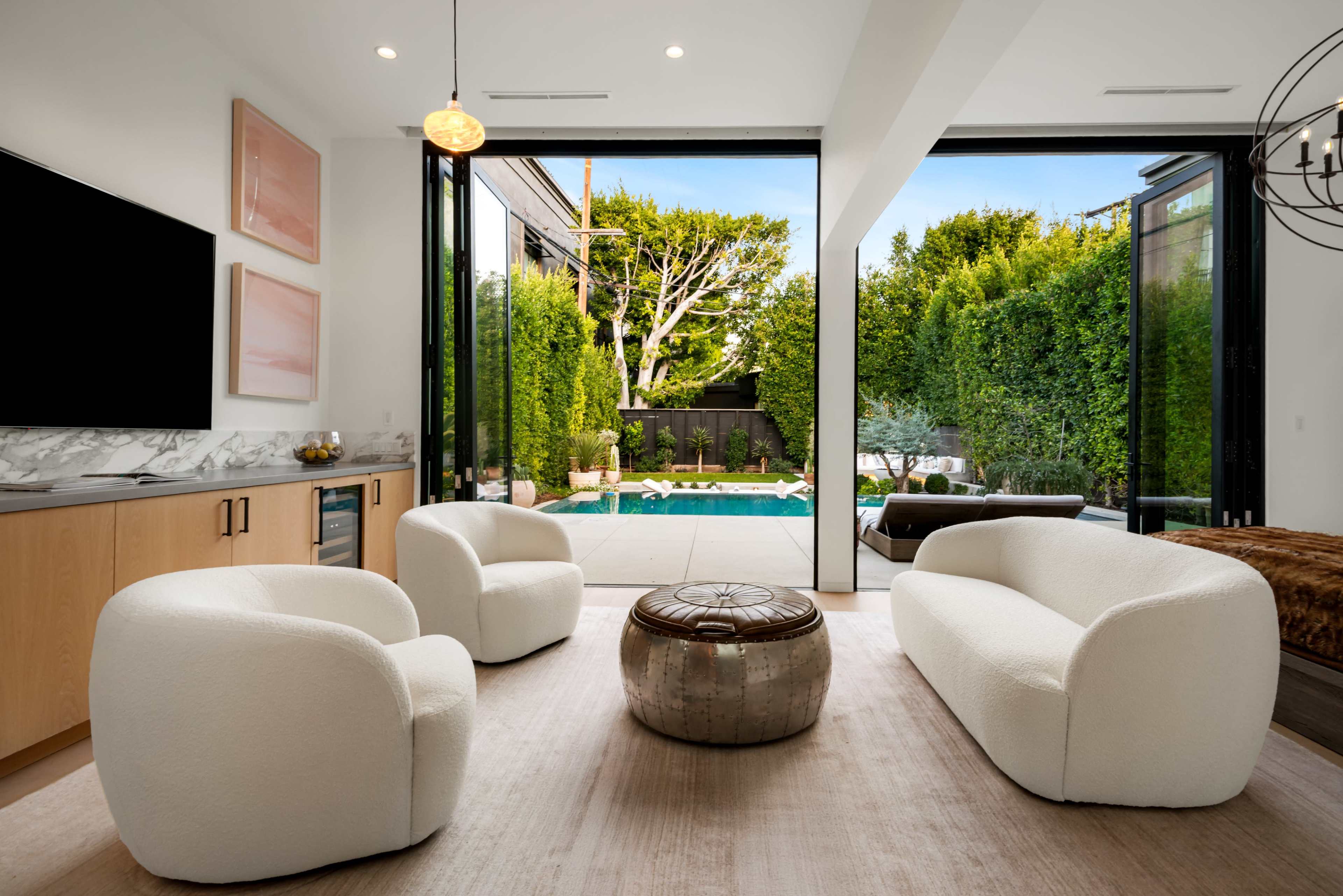 A modern living room features two white chairs and a round metallic table, with large glass doors opening to a lush outdoor area and swimming pool.