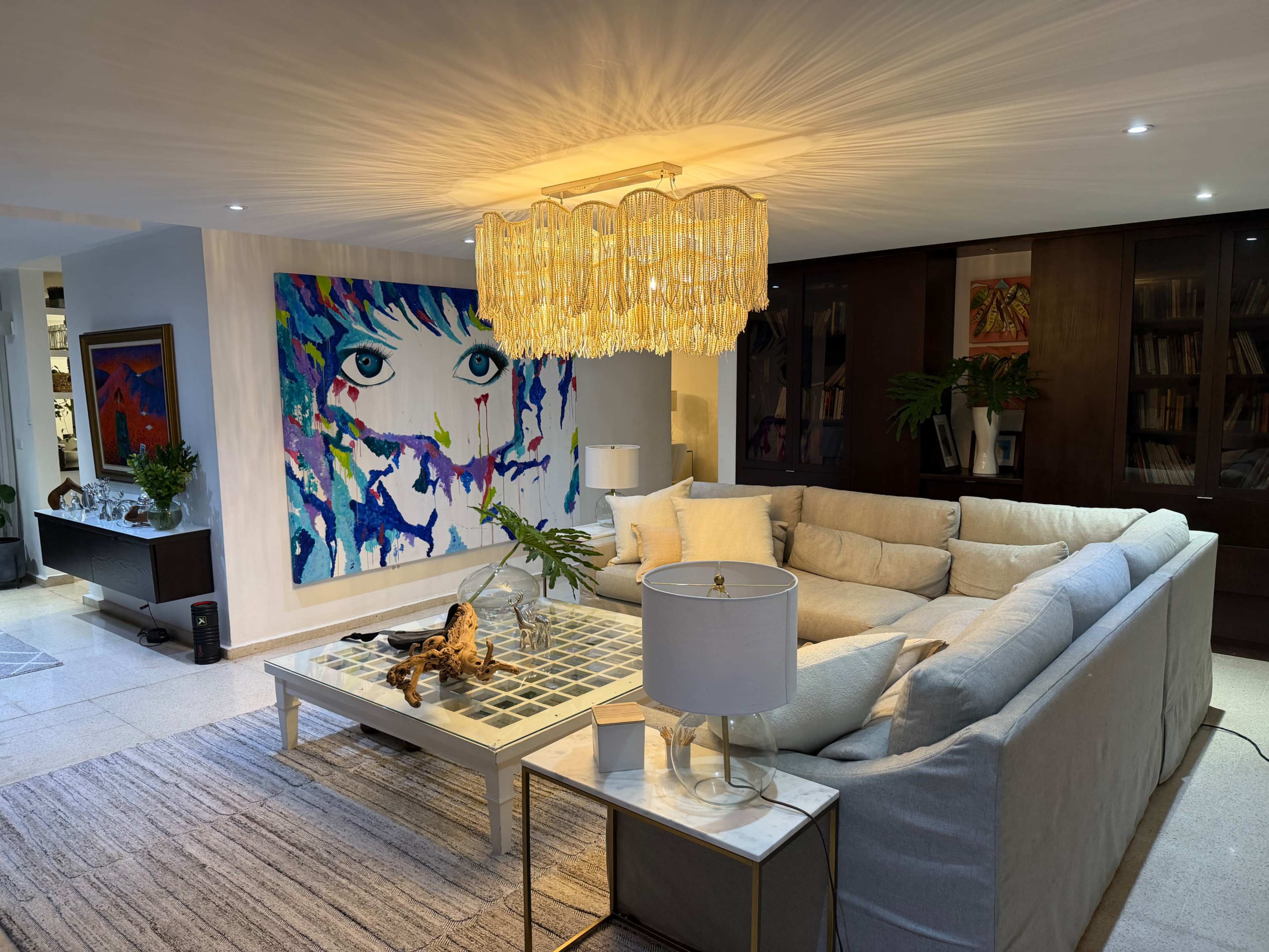 The image shows a modern living room featuring a large sectional sofa, a decorative coffee table, an eye-catching wall art piece, and a stylish chandelier.