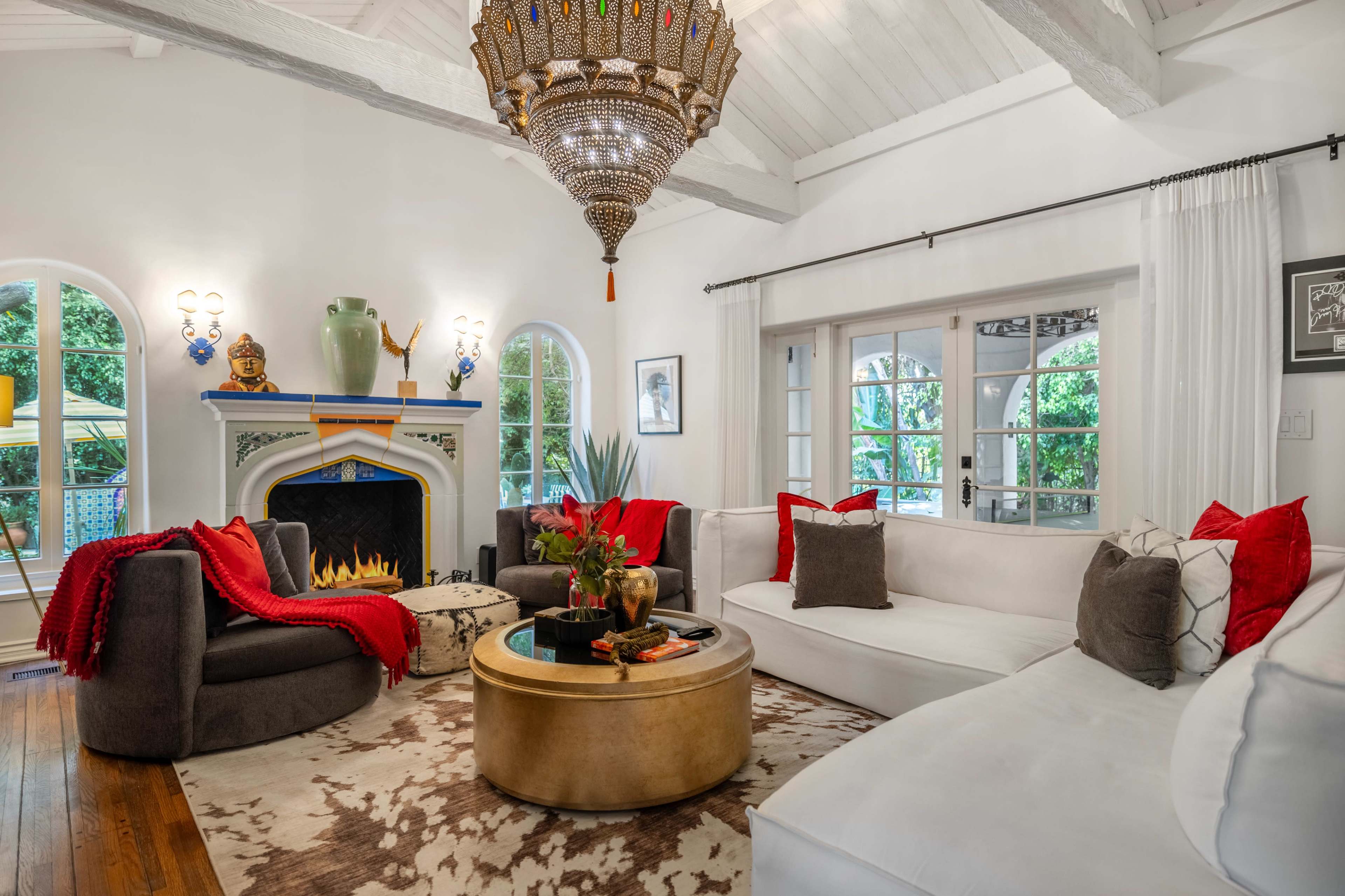 A cozy living room features a large white sectional sofa, a round gold coffee table, and a decorative fireplace, with various plants and colorful accents throughout the space.