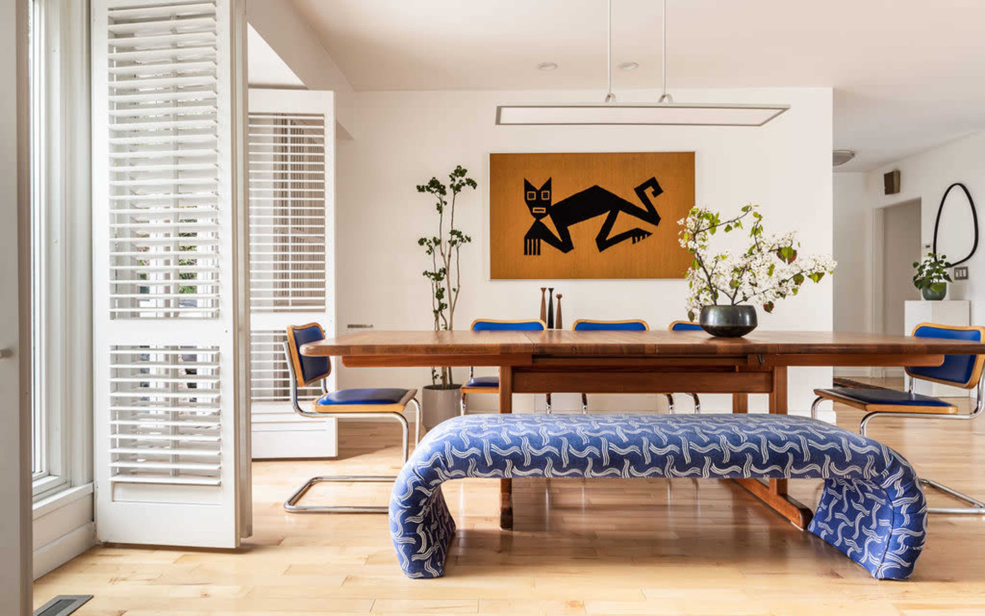 The image shows a modern dining area featuring a large wooden table, a blue and white patterned bench, and a wall adorned with a decorative artwork.
