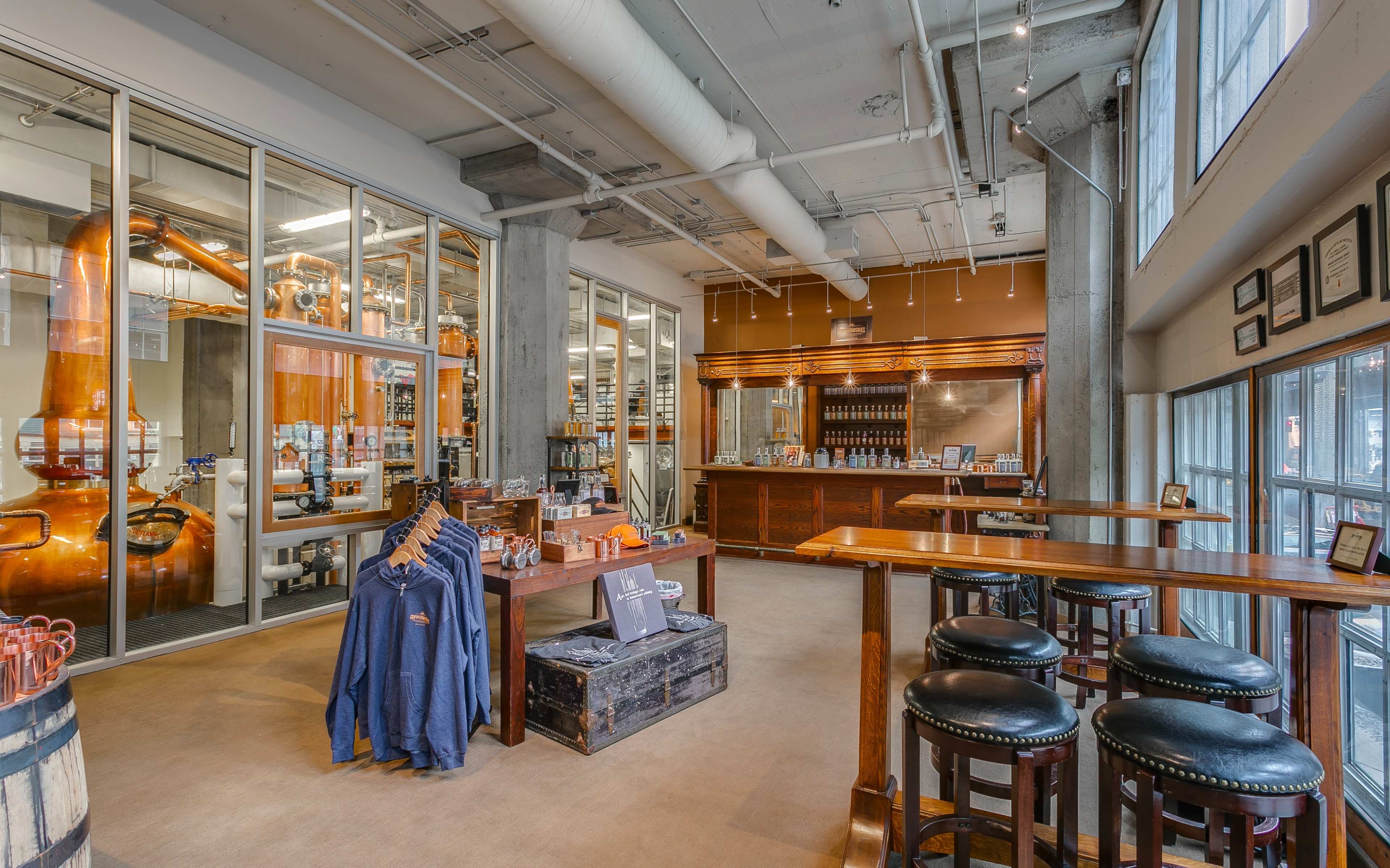 A spacious distillery bar area with large windows, a wooden counter, stools, and visible distillation equipment in the background.