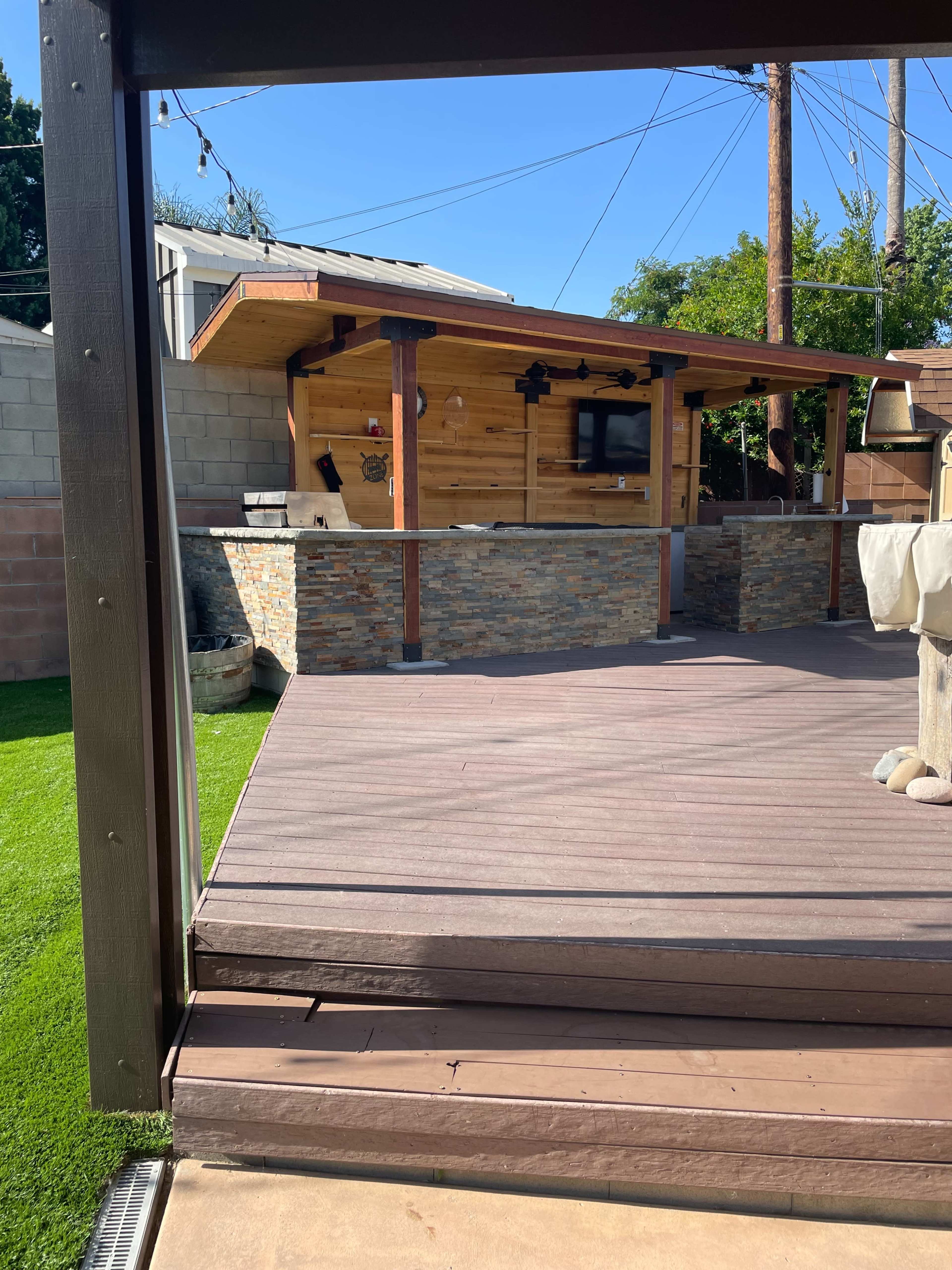 A wooden deck leading to an outdoor bar area made of stone and wood, surrounded by green grass and utility poles in the background.