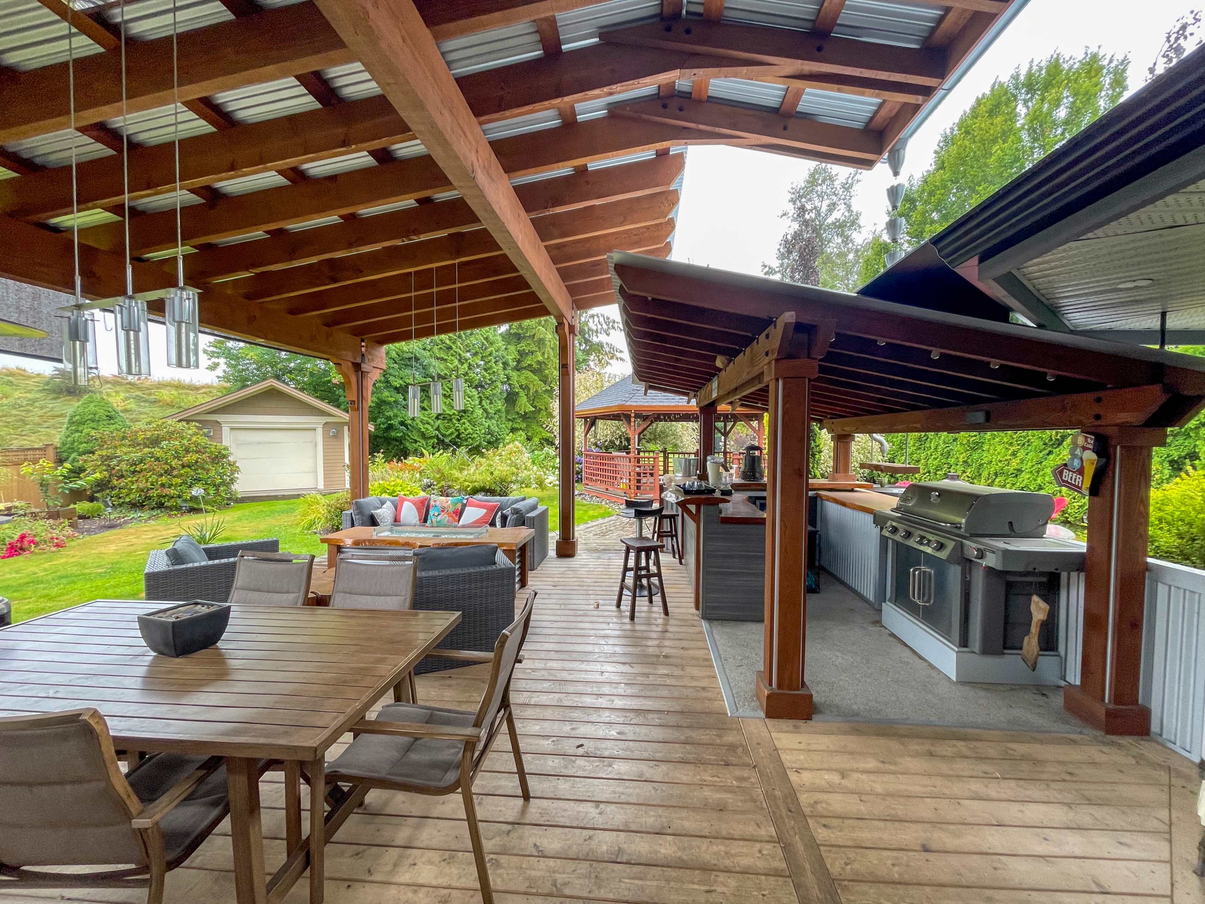 A covered outdoor patio area with a dining table, lounge seating, and a barbecue grill setup, surrounded by greenery.
