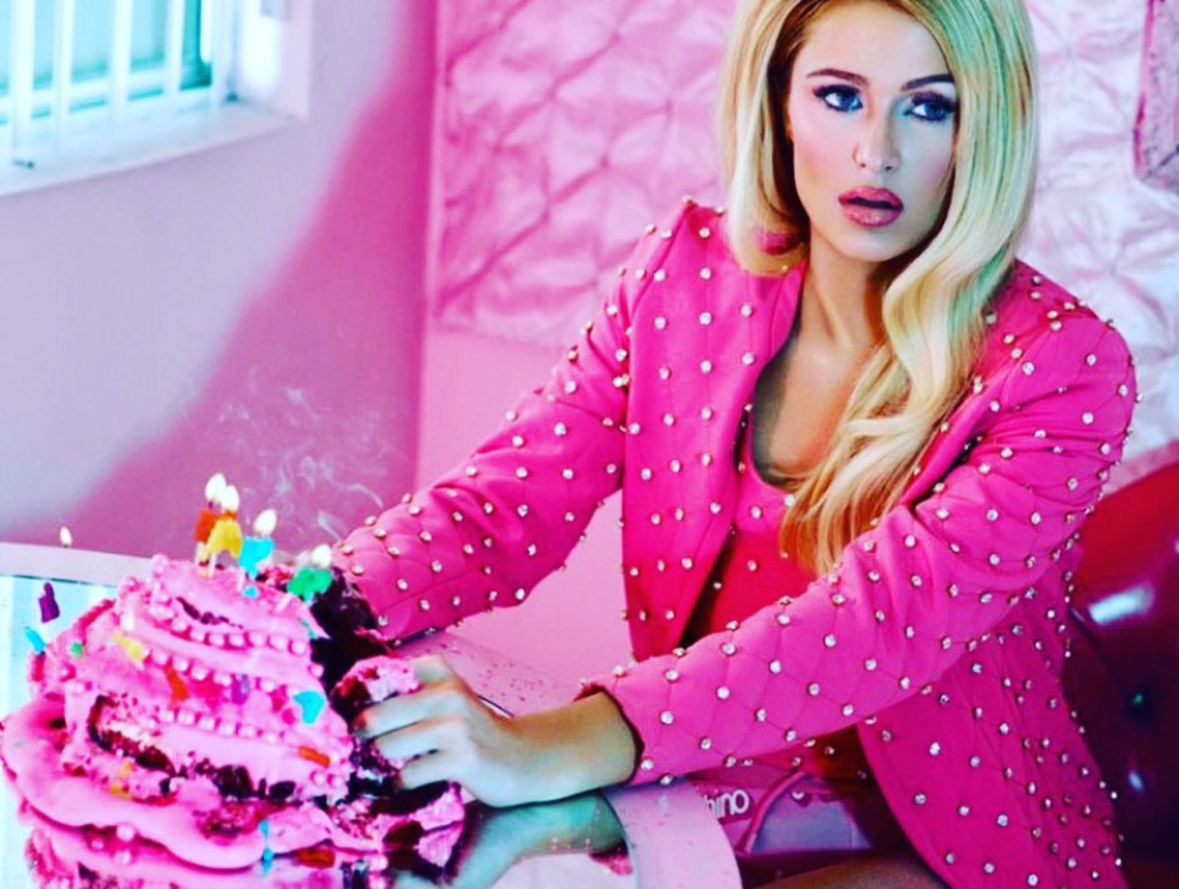 A person with long blonde hair sits at a table in a pink room, next to a decorated pink birthday cake with candles.