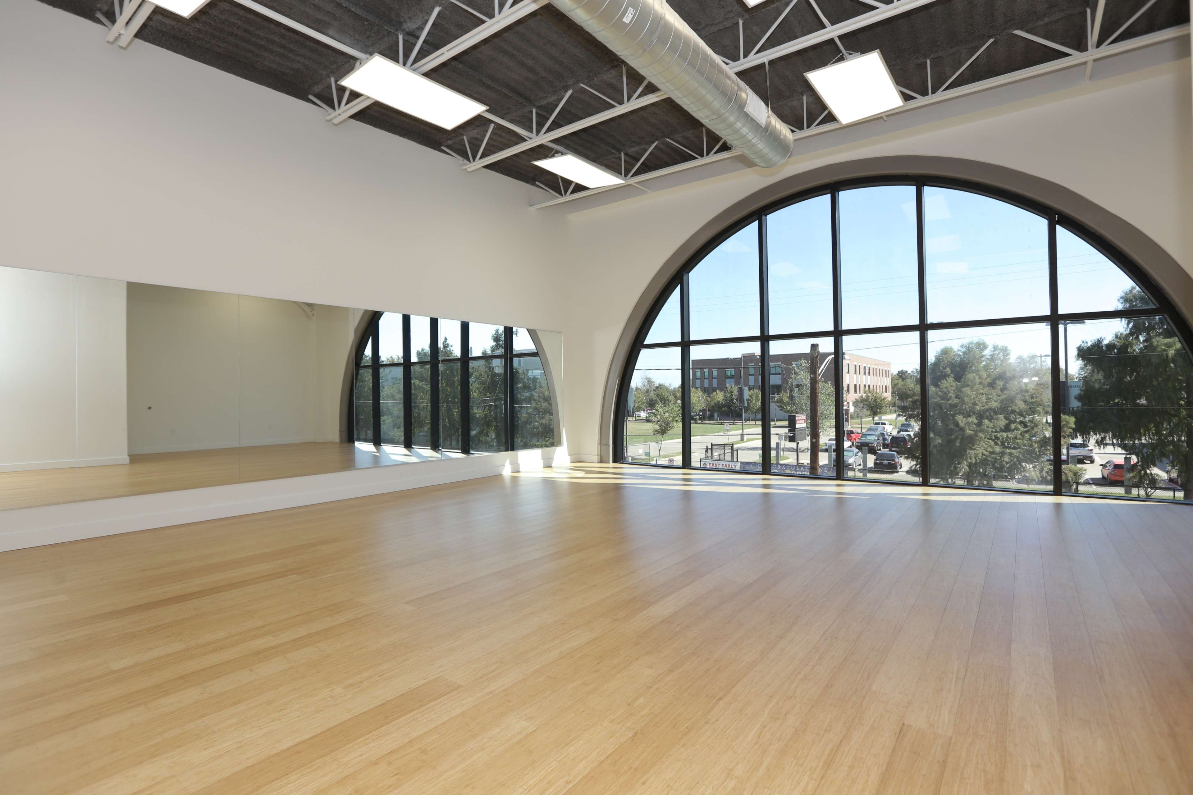 A large, well-lit dance studio features a curved window, a mirror along one wall, and a bamboo floor.