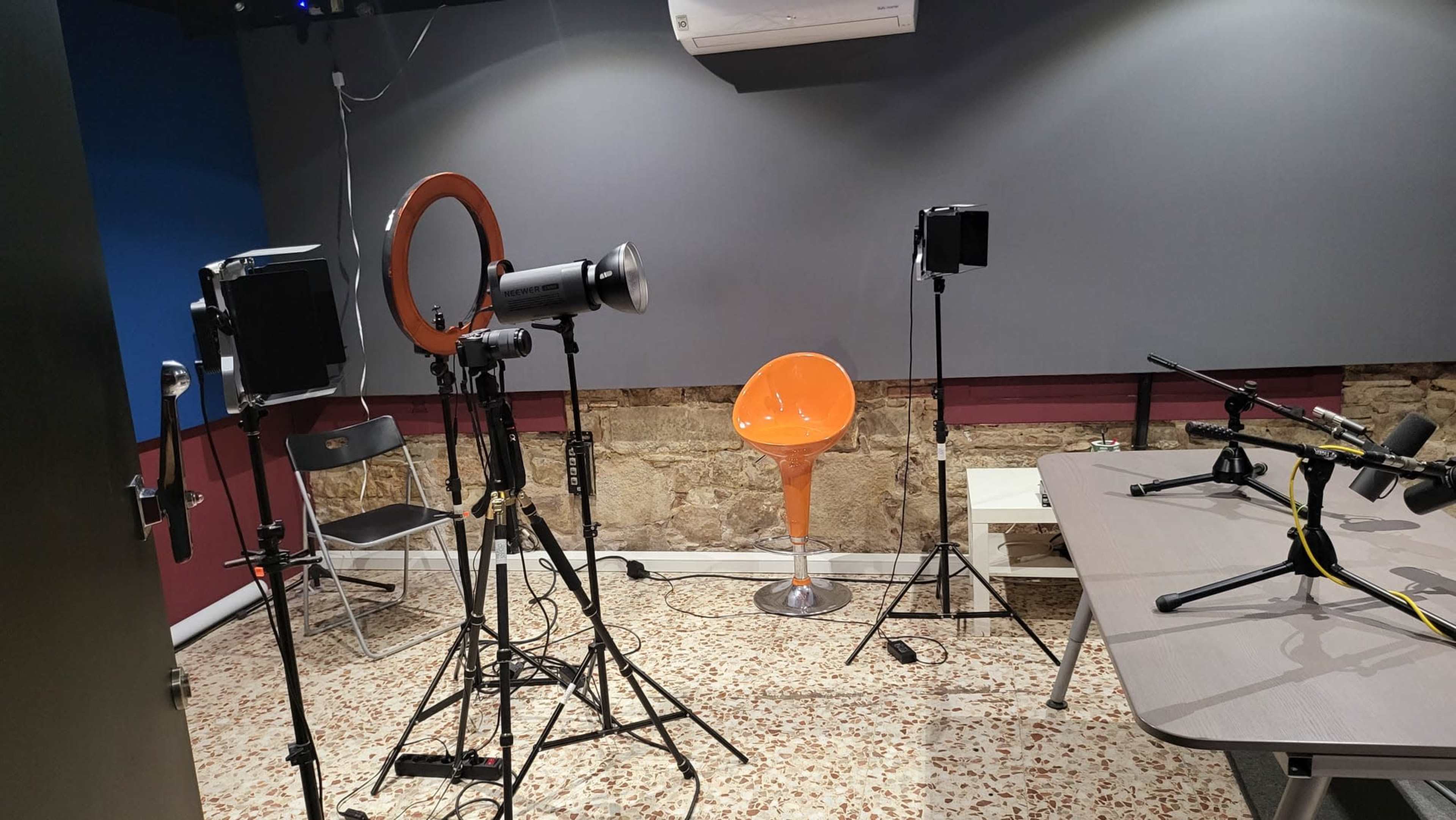 A studio setup with lighting equipment, an orange chair, a table with microphones, and a textured wall in the background.