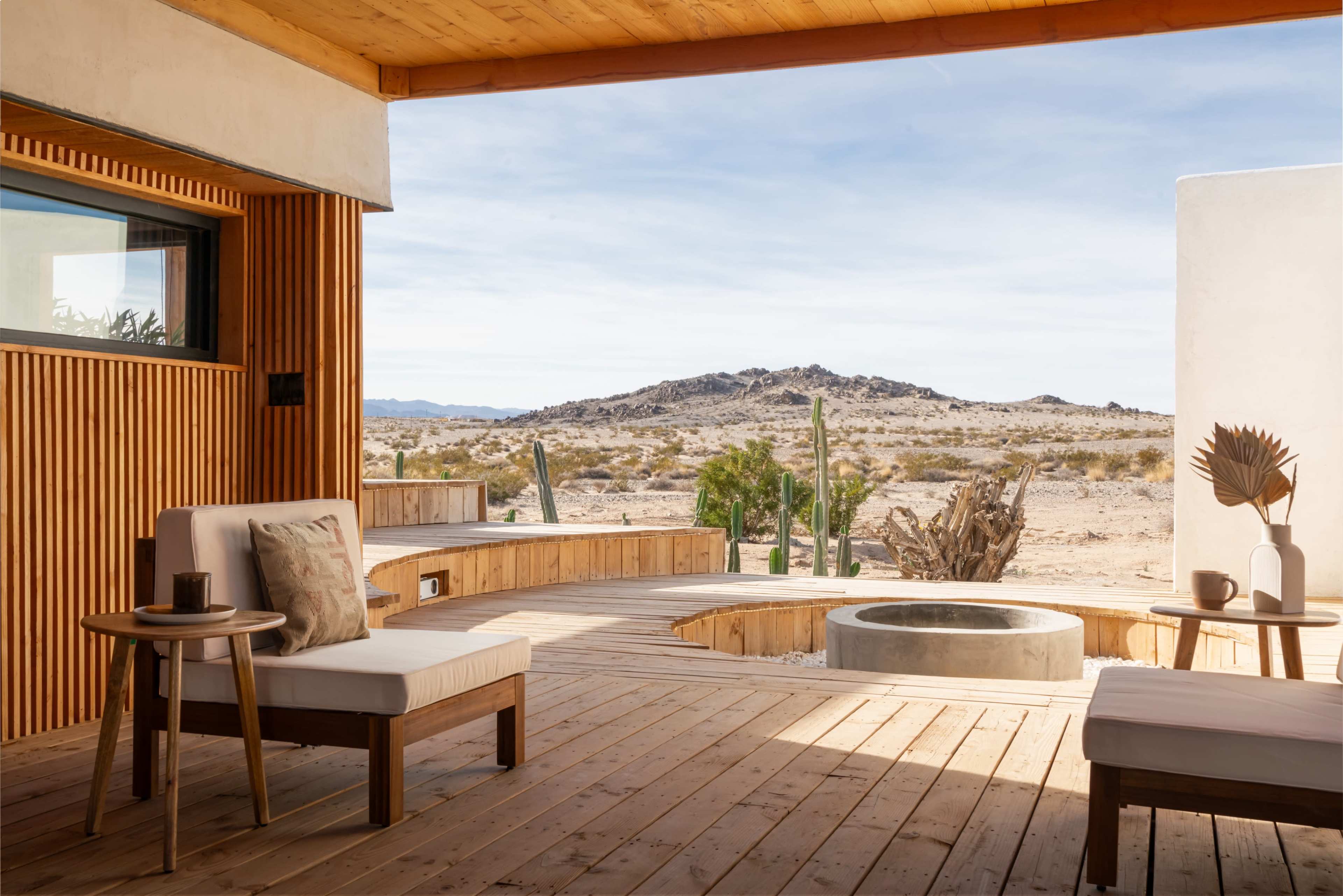 The image shows a modern, wooden patio with minimalistic seating and a circular fire pit, overlooking a vast desert landscape with rocky hills and sparse vegetation.