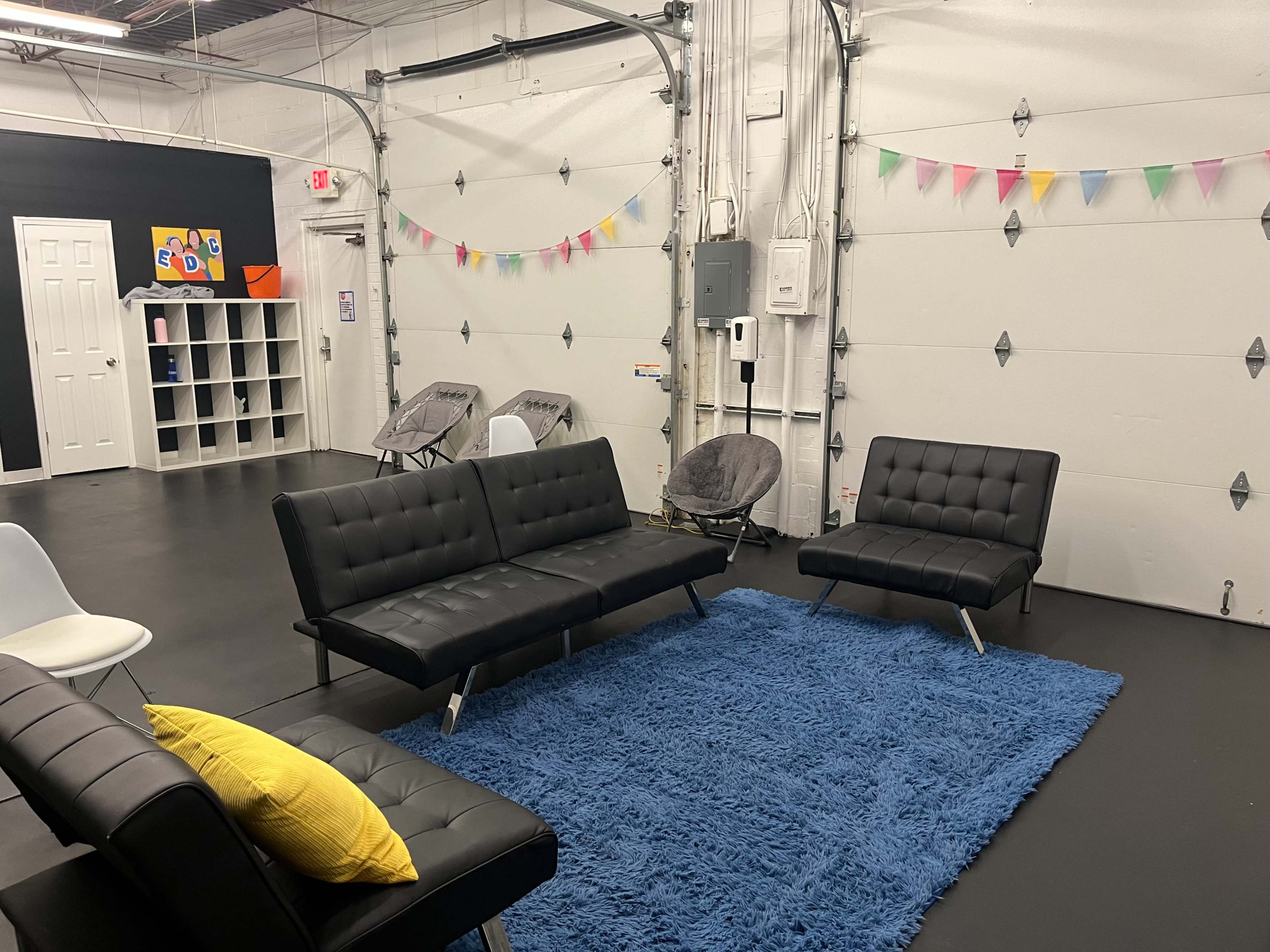 A modern, minimalistic lounge area with black sofas, a blue rug, and colorful decorations strung along the walls.