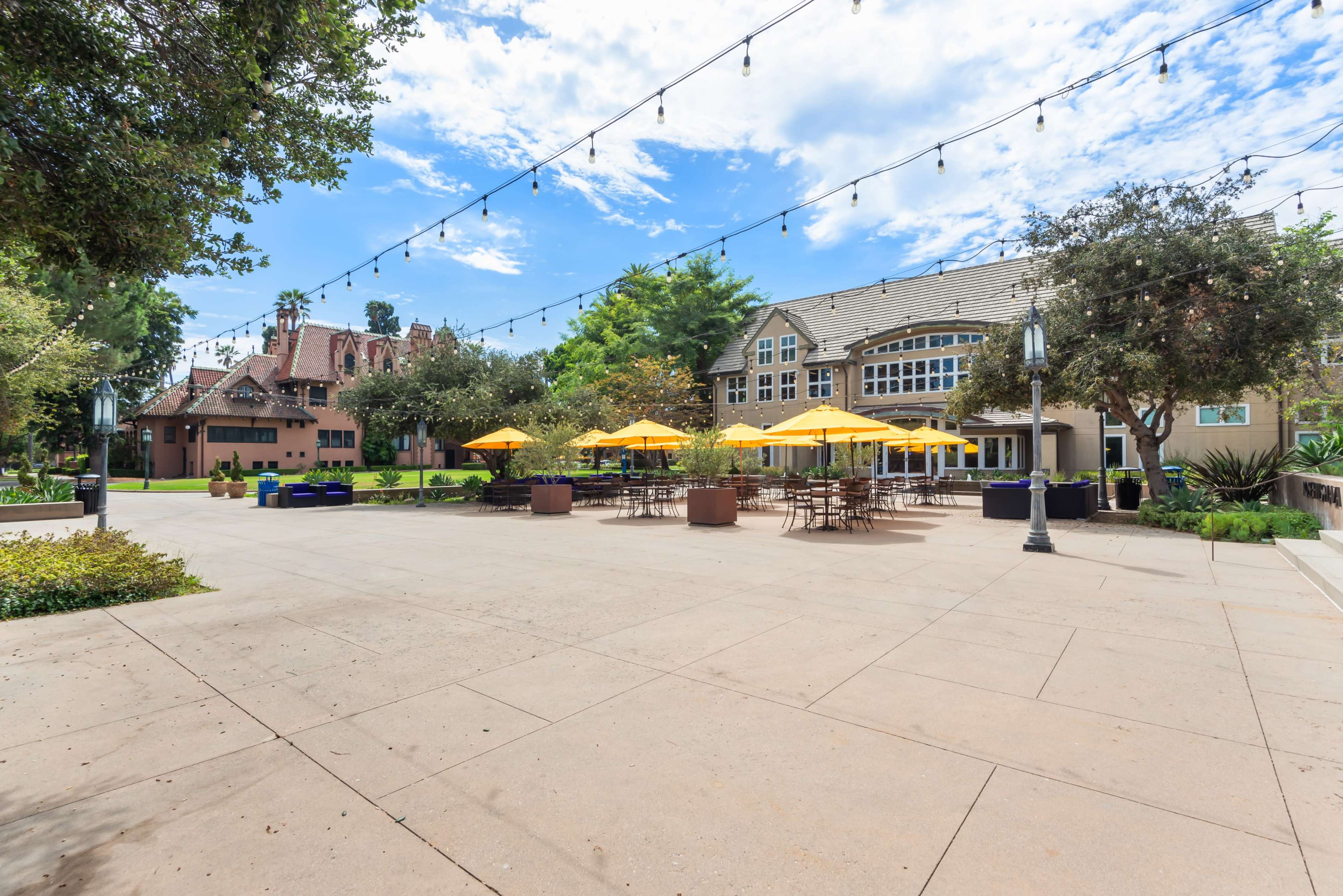 The image shows a paved outdoor area with yellow umbrellas over tables, surrounded by trees and a large building in the background.