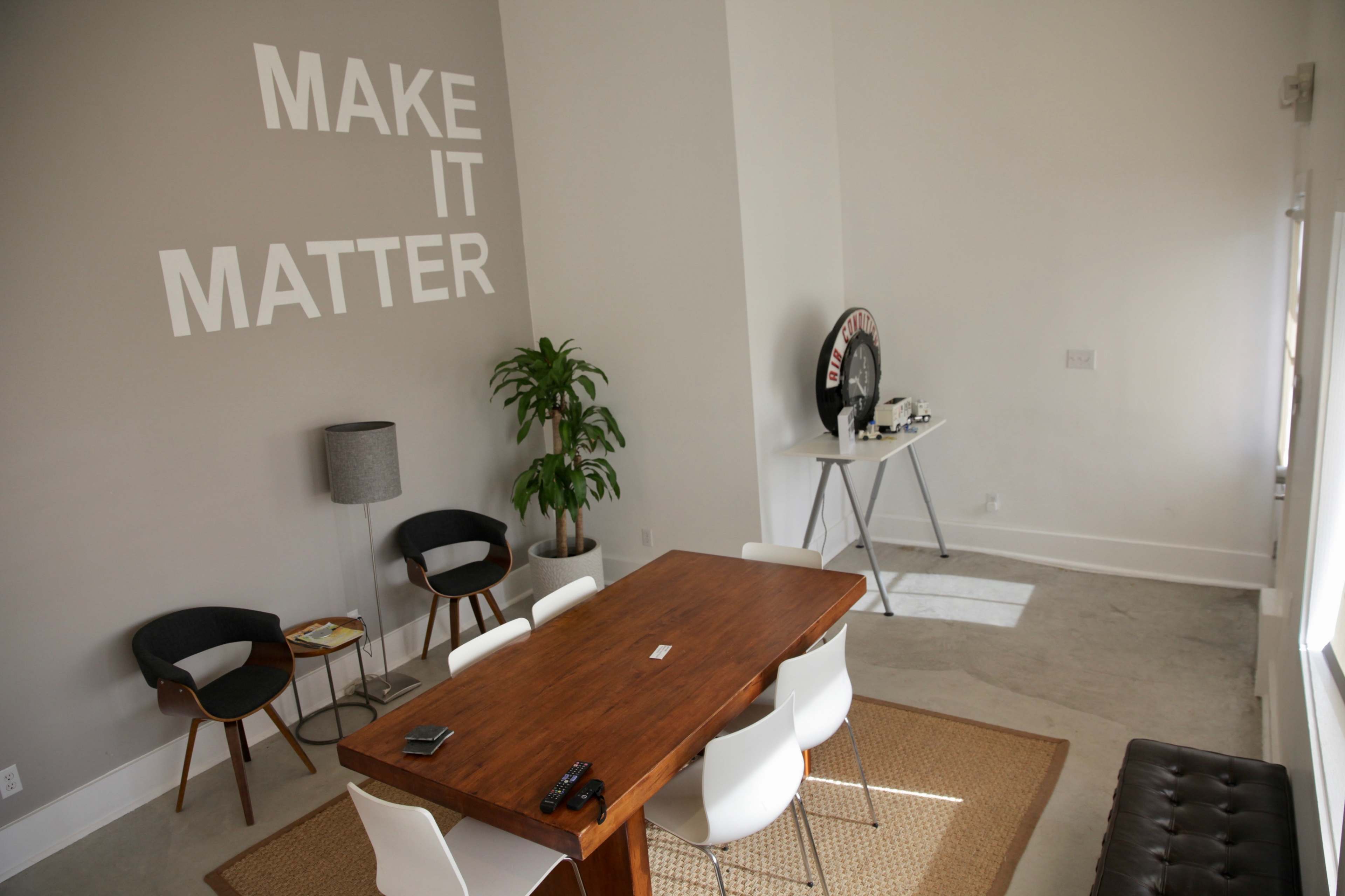 A spacious room features a wooden dining table surrounded by white chairs, a potted plant in the corner, and the phrase "MAKE IT MATTER" written on the wall.