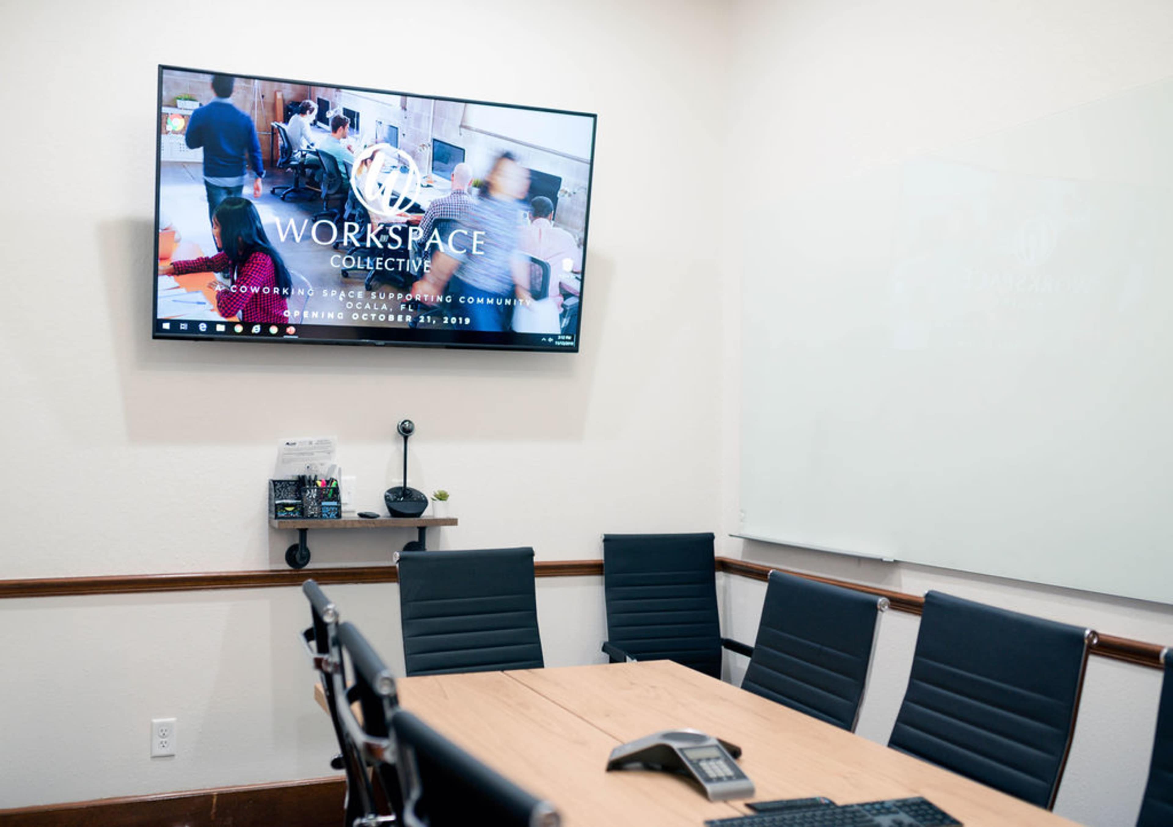A meeting room with a large wall-mounted screen displaying "Workspace Collective" and a table surrounded by black office chairs.