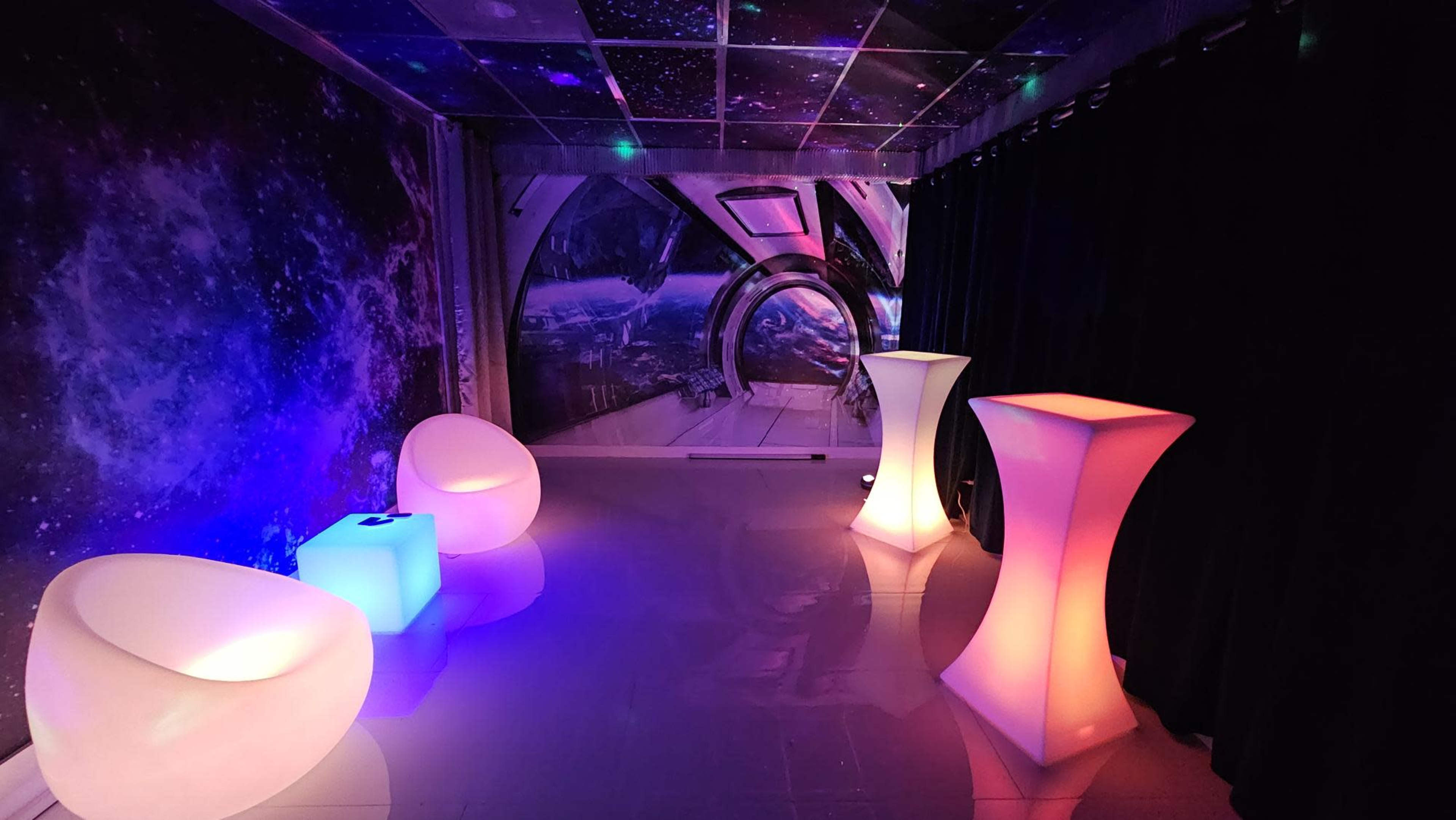 A room decorated with a cosmic mural and illuminated furniture, including two glowing tables and two curved chairs, set against dark curtains.