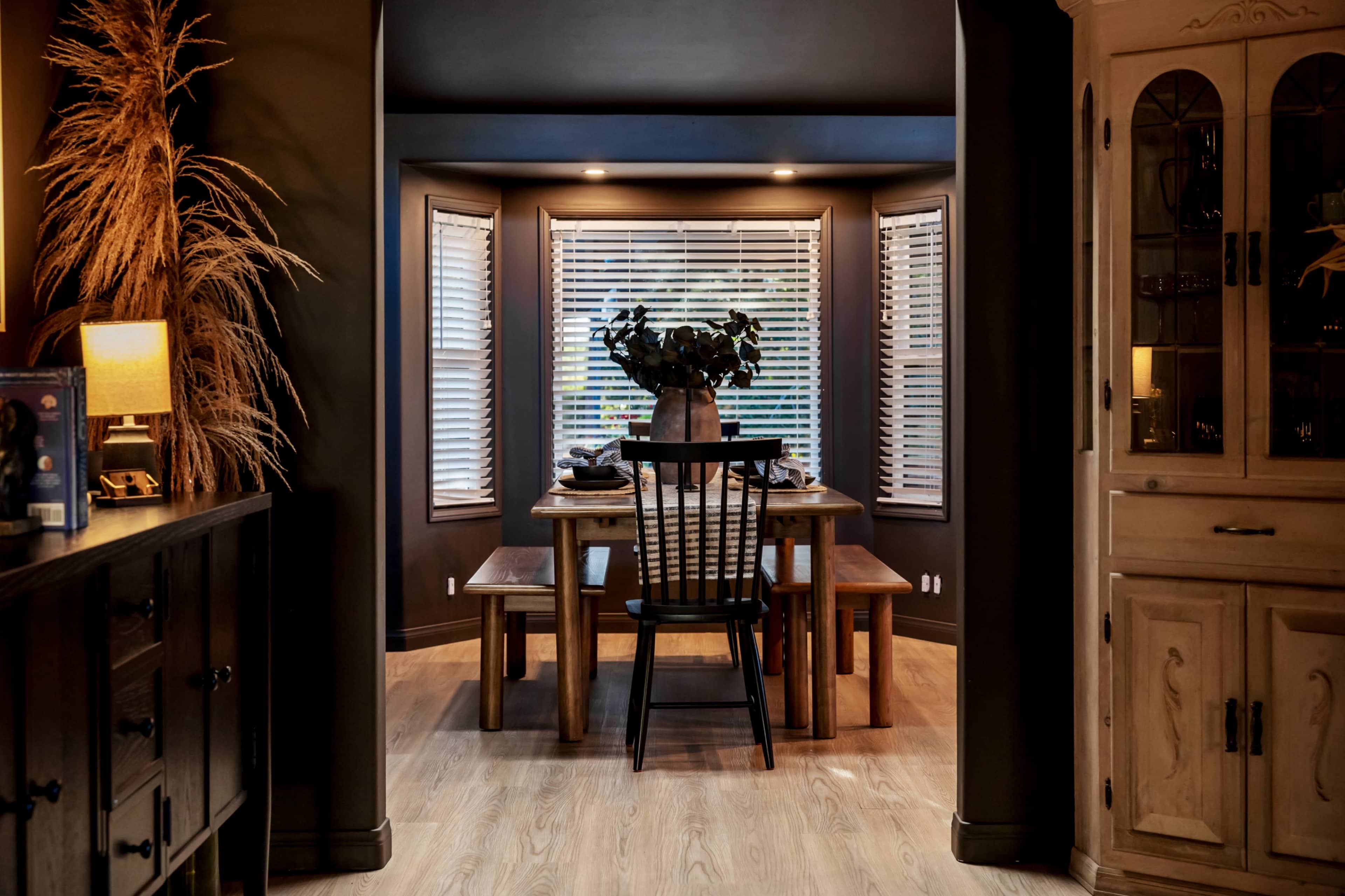 A wooden dining table with two chairs is set in a corner nook adorned with windows, surrounded by dark walls and a cabinet.