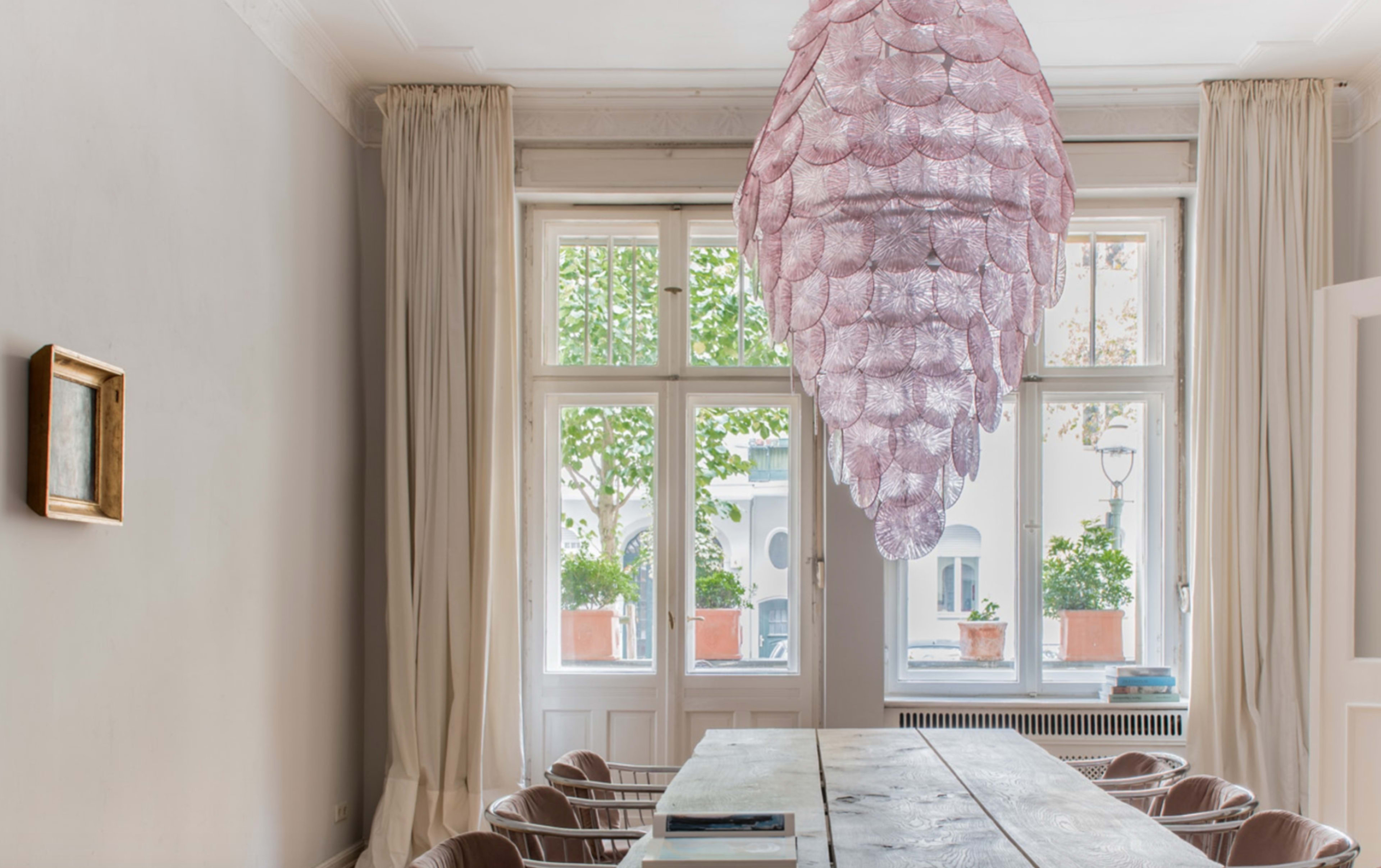 A dining room features a large, pink light fixture hanging above a wooden table, with large windows allowing natural light and greenery from outside.