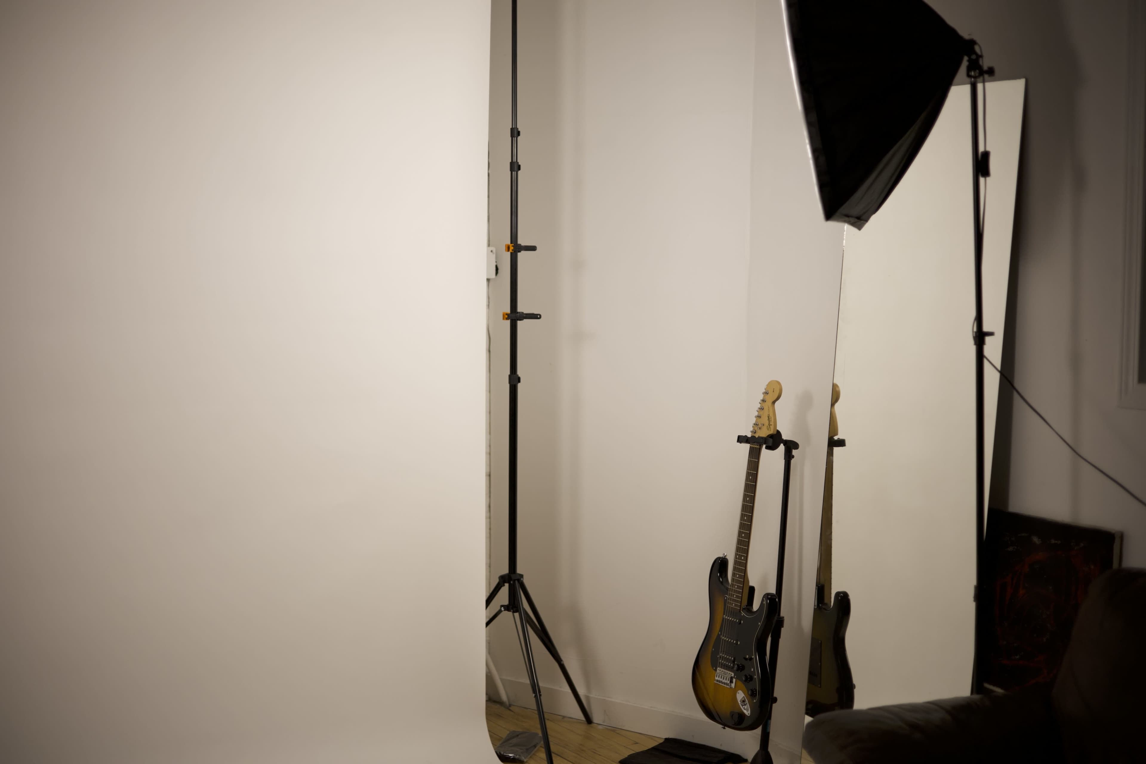 The image shows a photography studio setup featuring a plain white backdrop, a lighting umbrella, a light stand, and two electric guitars leaning against a wall.